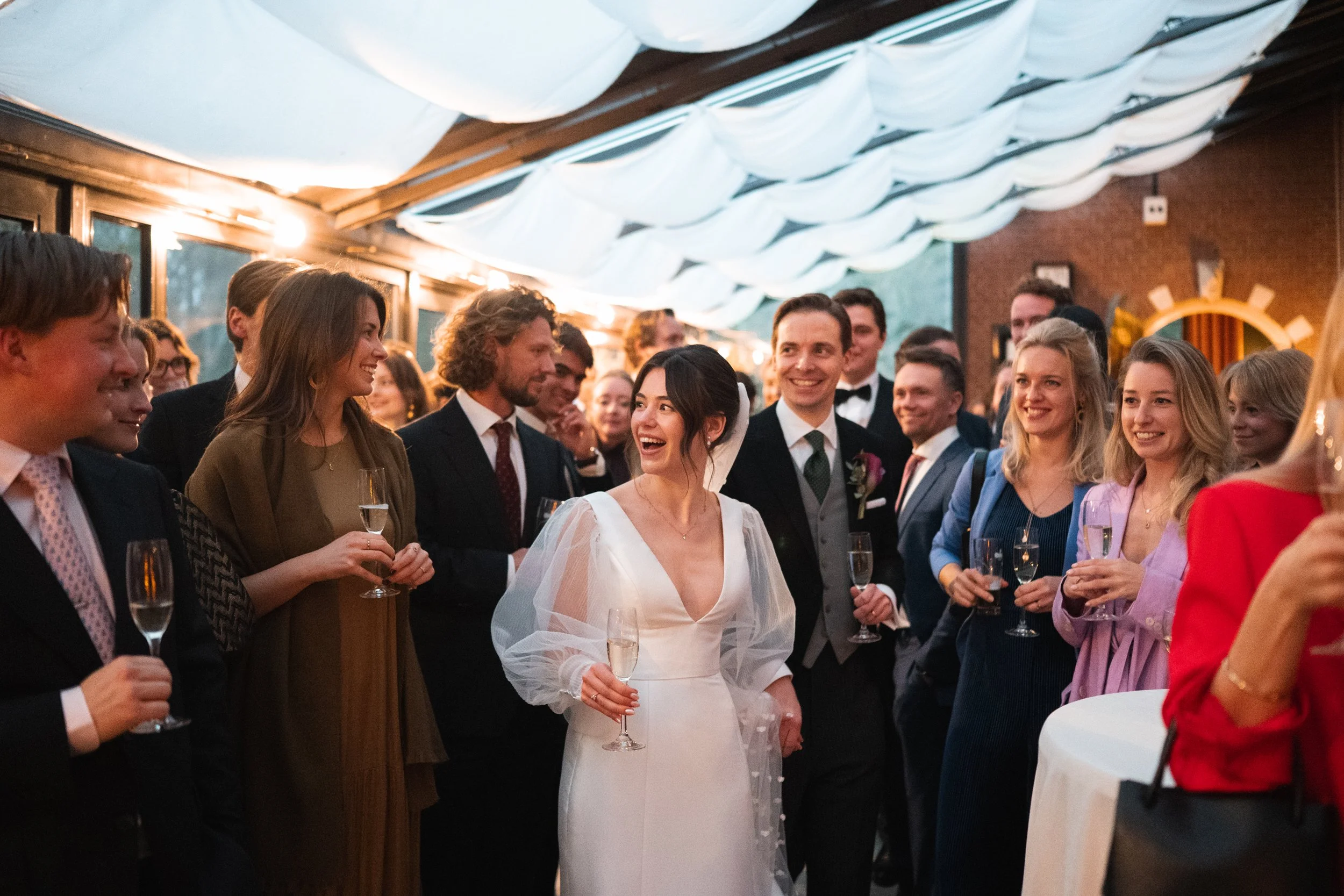 Wedding reception with a bride and groom smiling and holding champagne glasses, surrounded by friends and family in an indoor venue.