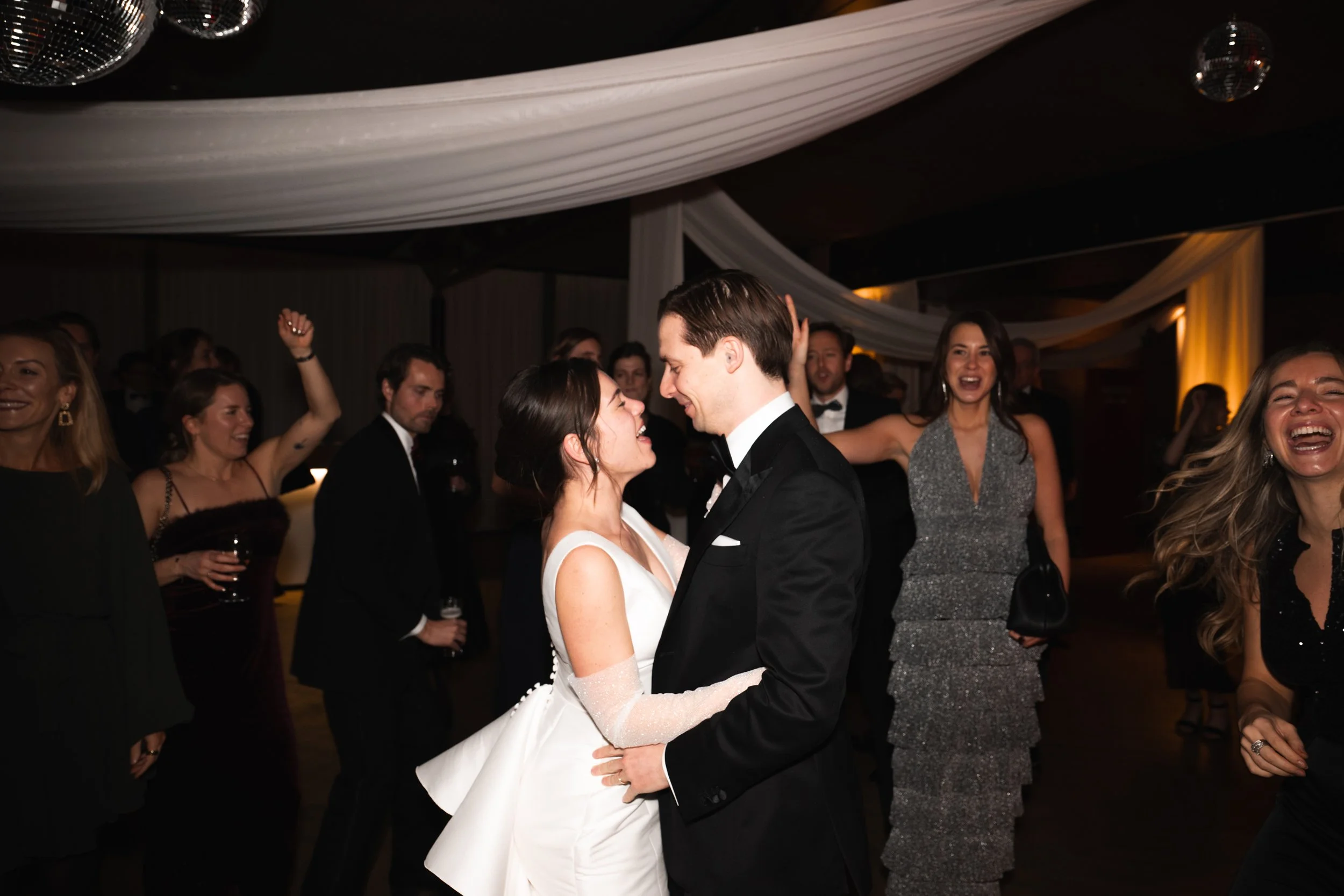 A bride and groom dance closely at their wedding reception, surrounded by laughing and celebrating guests.