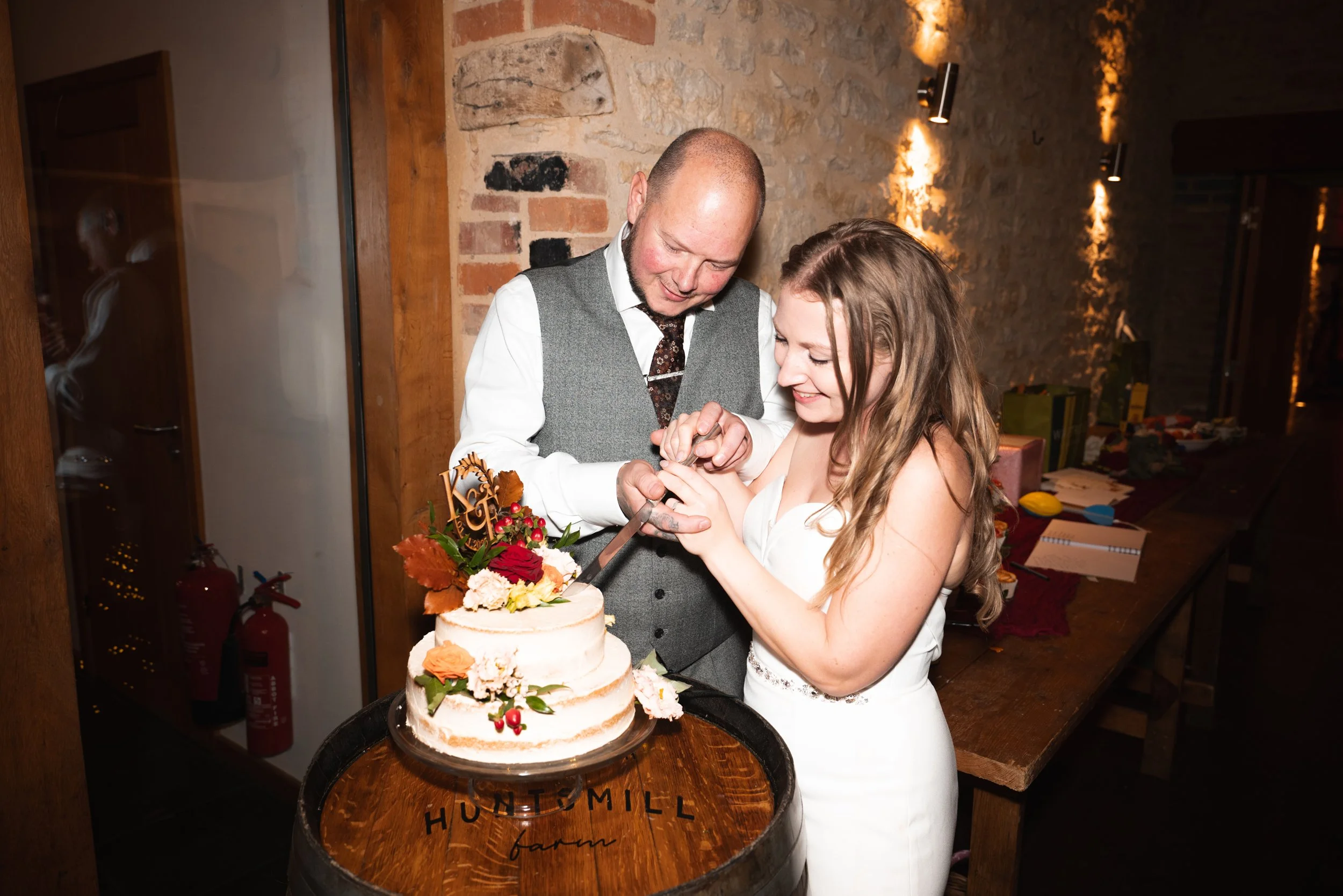 A bride and groom cutting their wedding cake together during a wedding reception in a rustic venue with brick walls.