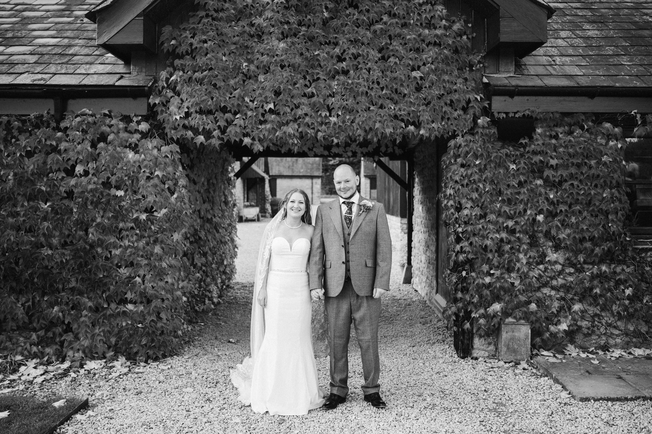 A bride and groom holding hands and smiling in front of a rustic stone archway with ivy, outdoors during daytime.