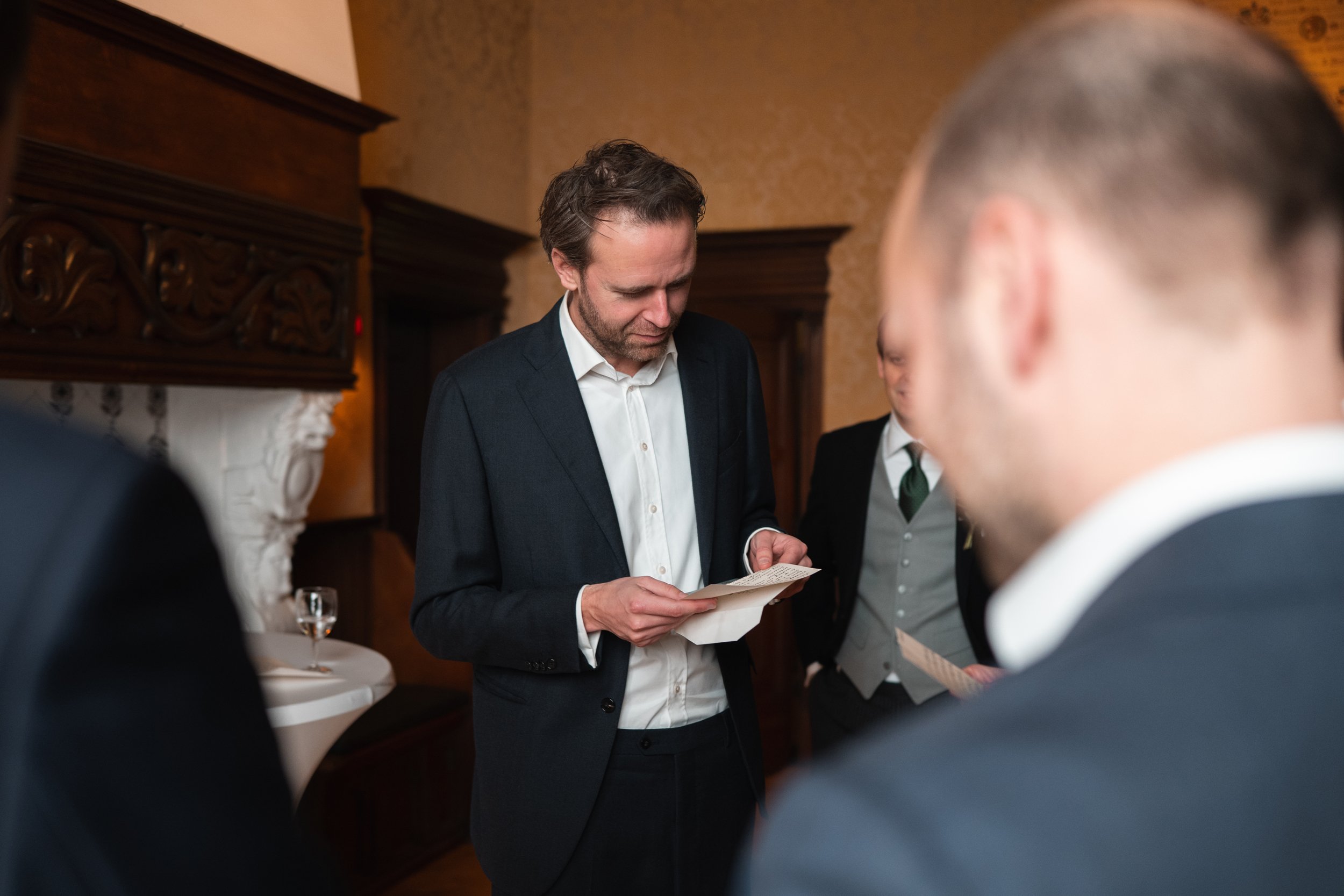 A man in formal attire looking at a printed document in a room with wood-paneling and ornate white fireplace.
