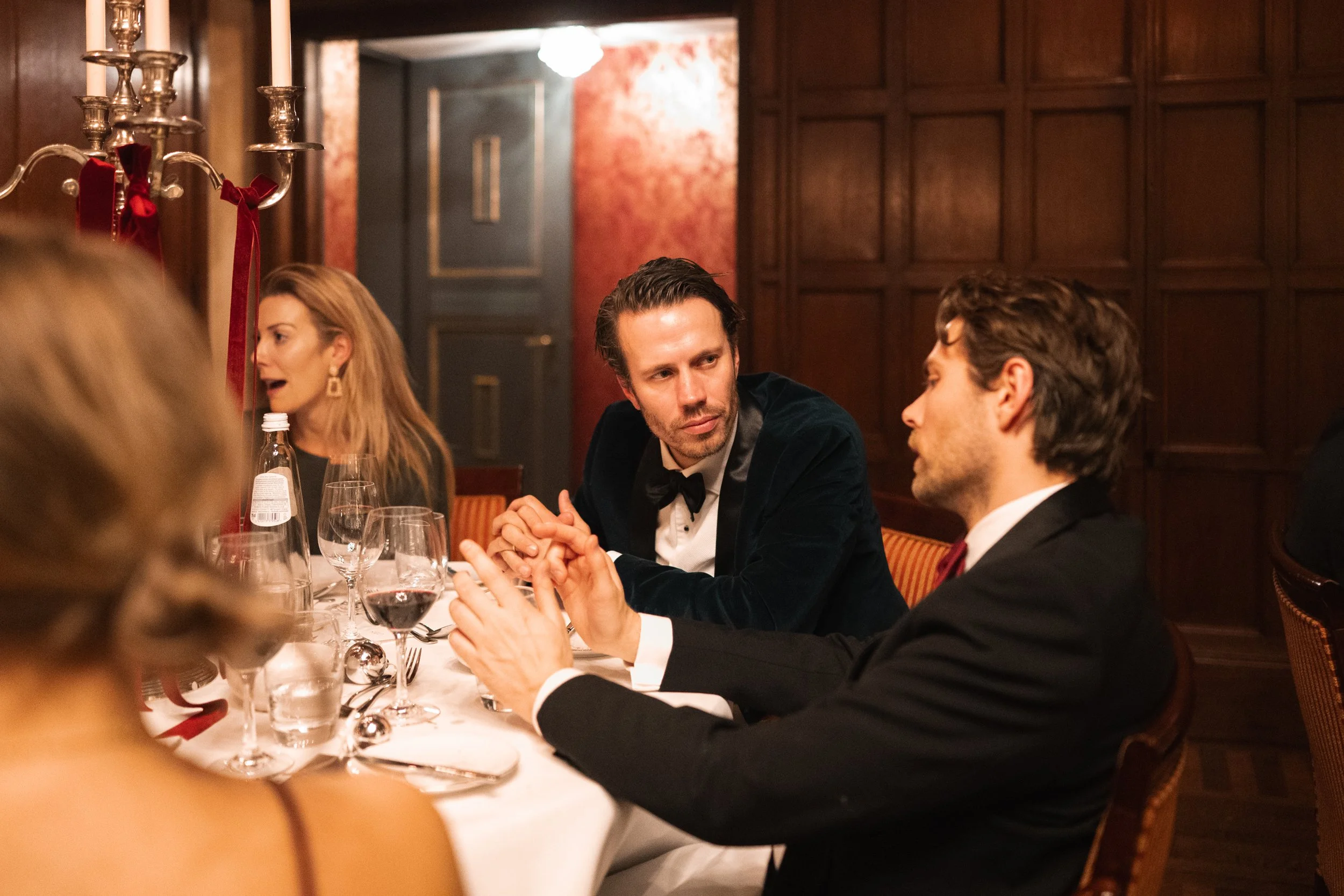 A group of well-dressed people having a conversation at a dinner party in an elegant, wood-paneled room with a candelabra on the table.