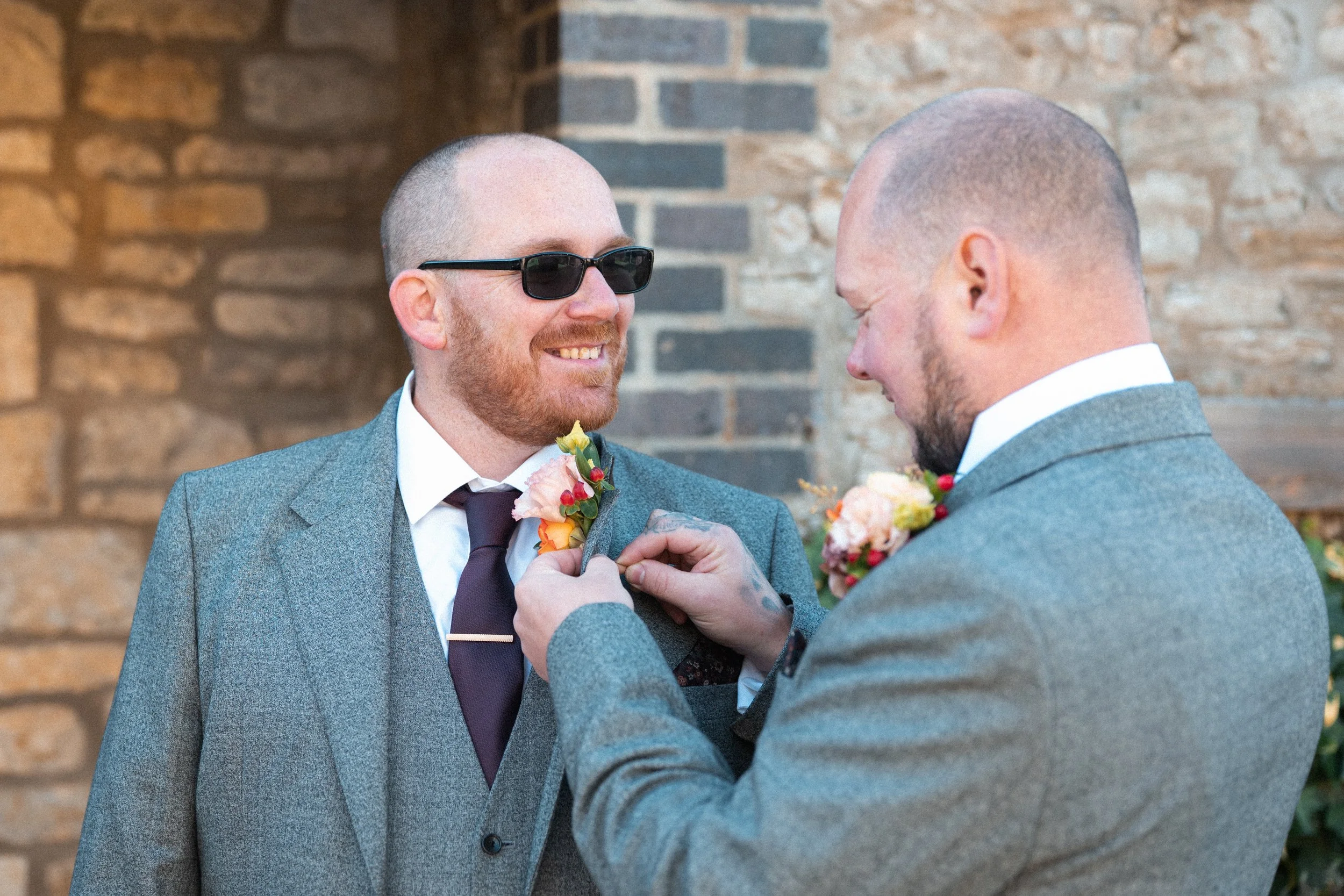 Two men in suits celebrating a wedding, one wearing sunglasses, as the other pins a boutonniere on his lapel in front of a brick wall.