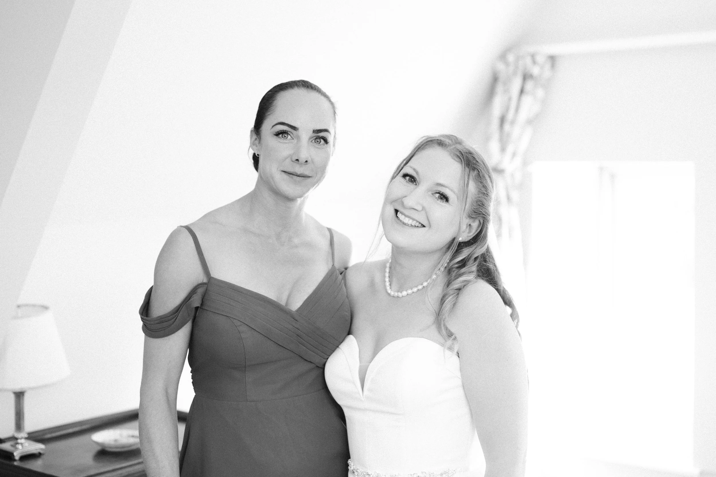 Black and white photo of a woman in a formal dress and a young girl in a wedding gown, smiling in a bright room with a window and curtains.