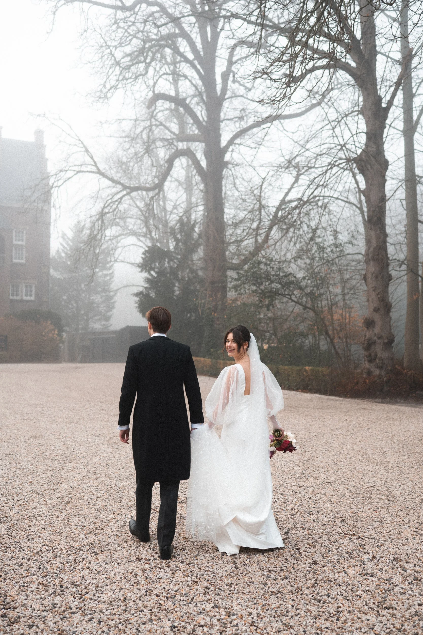 Bride and groom walk hand in hand outdoors on a gravel path, with foggy trees and a building in the background. The bride is smiling and holding a bouquet, wearing a white wedding dress with sheer sleeves. The groom wears a black tuxedo, with his bac