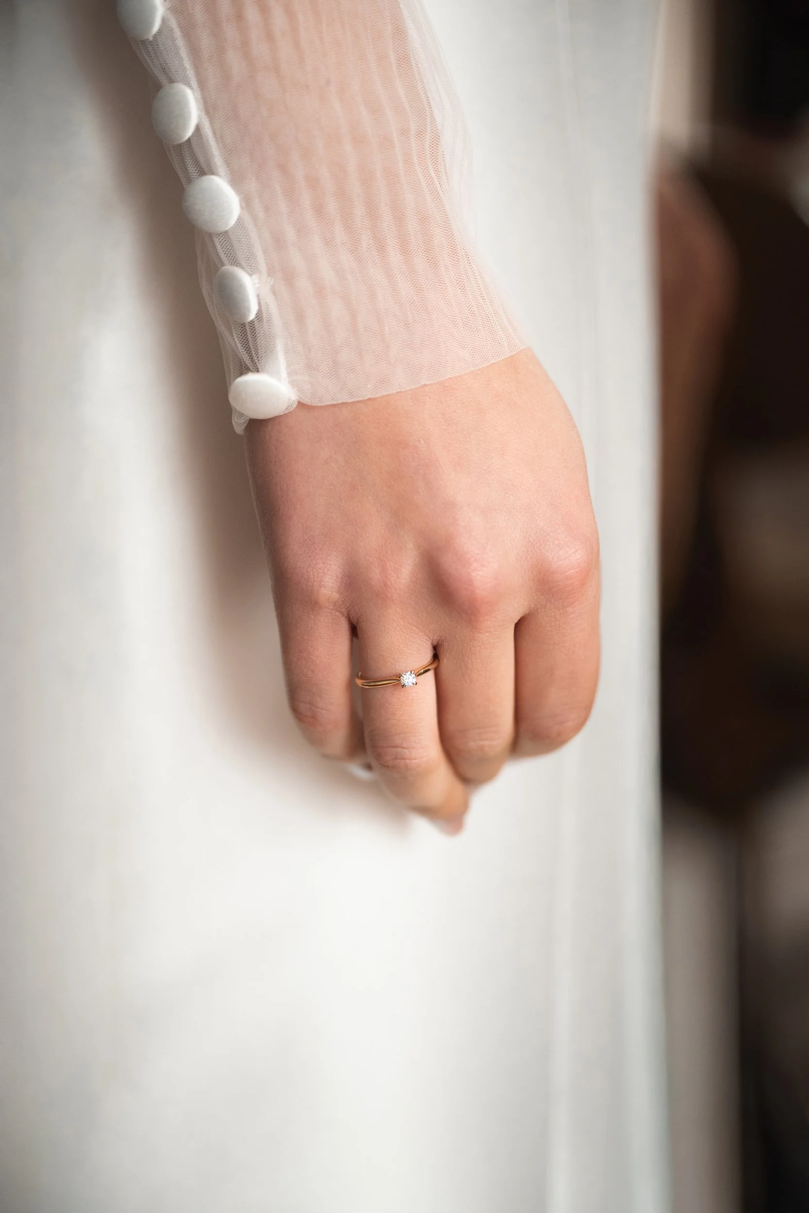 Close-up of a woman's hand resting on a white surface, wearing a gold ring with a small gemstone on her ring finger, and a light-colored, sheer sleeve with button details.