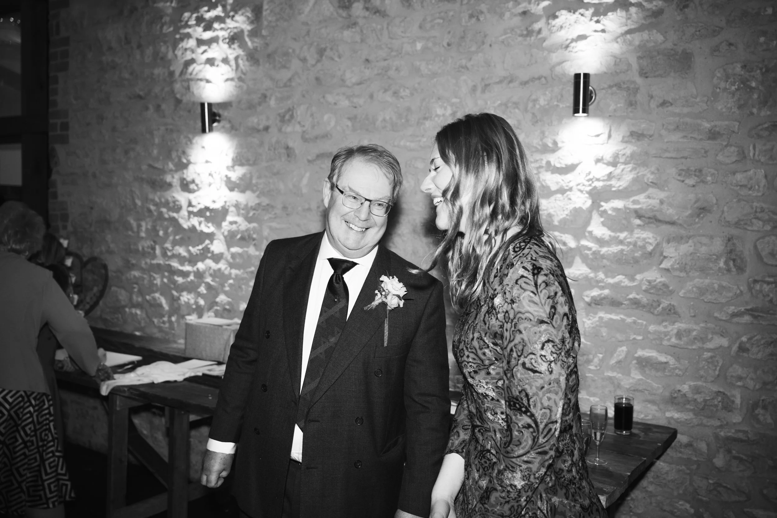 A smiling man in a suit and a woman with wavy hair smiling at each other at a social event in a room with stone walls and wall-mounted lights, with drinks on a table in the background.