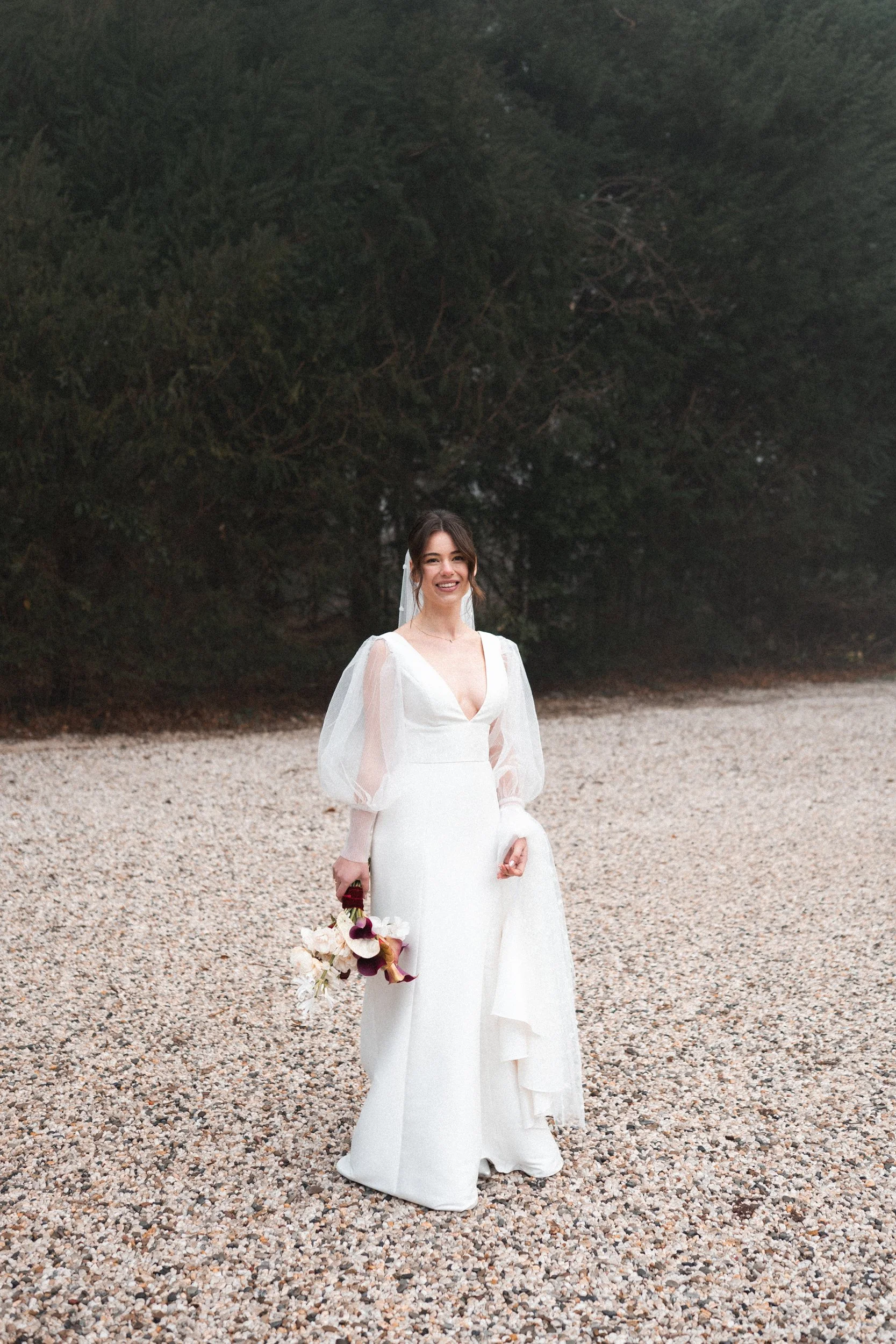 A bride standing outdoors on a gravel surface, holding a bouquet of flowers, smiling with dark hair and a white wedding gown with sheer puff sleeves, in front of a line of trees.