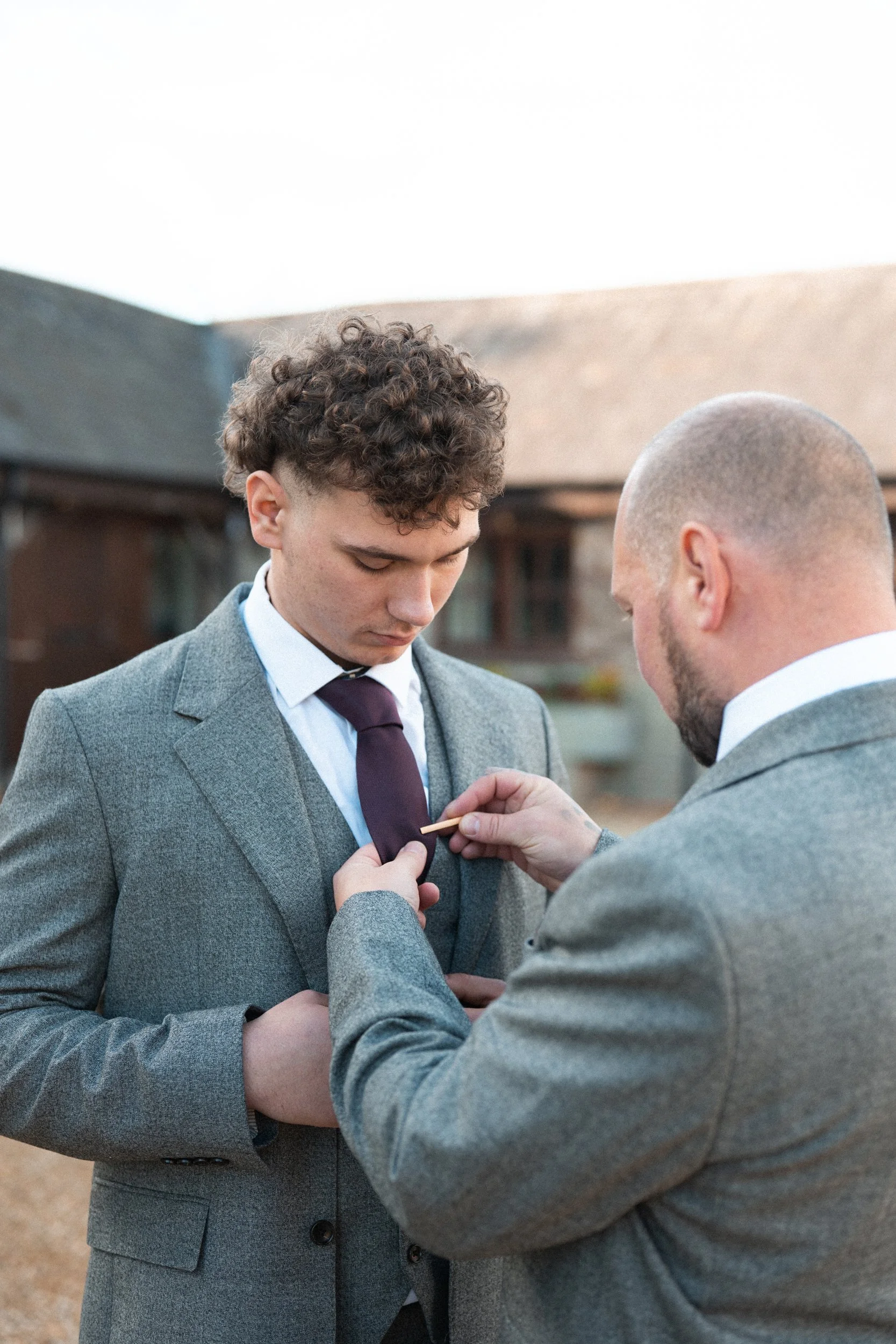 A man with curly hair being fitted with a boutonniere by another man with a shaved head, both wearing gray suits outdoors.