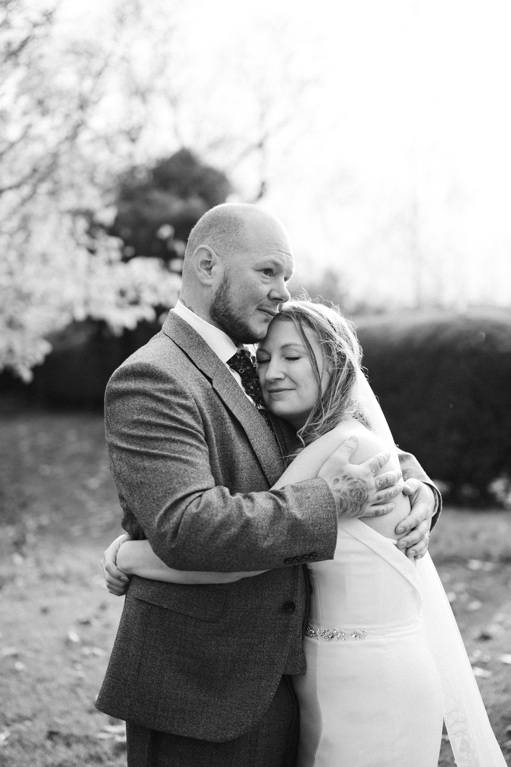 Black and white photo of a bride and groom hugging outdoors, with trees in the background.