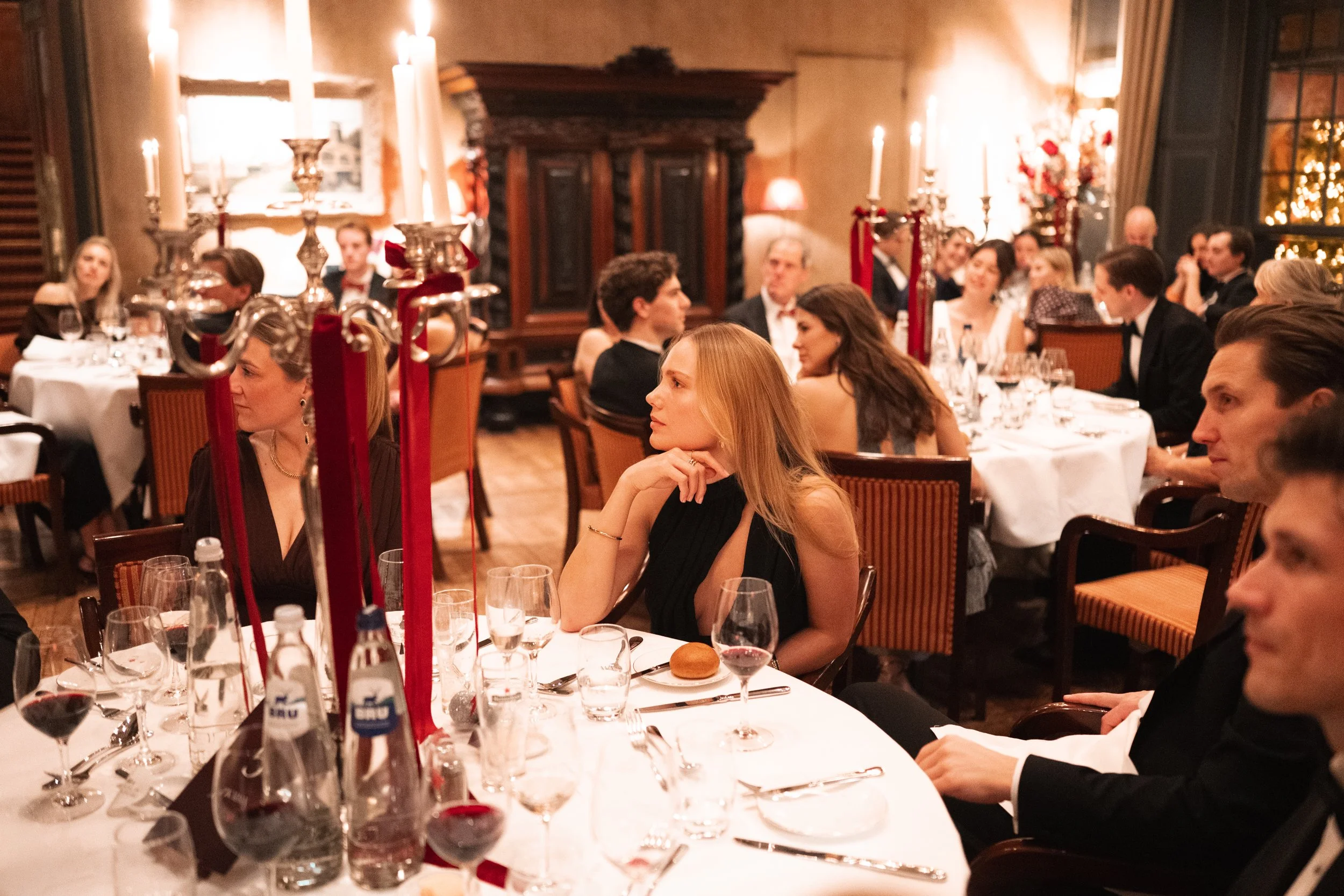 People seated around a round dining table at an elegant dinner party or formal event, with candles and wine glasses on the table in a warmly lit, decorated room.