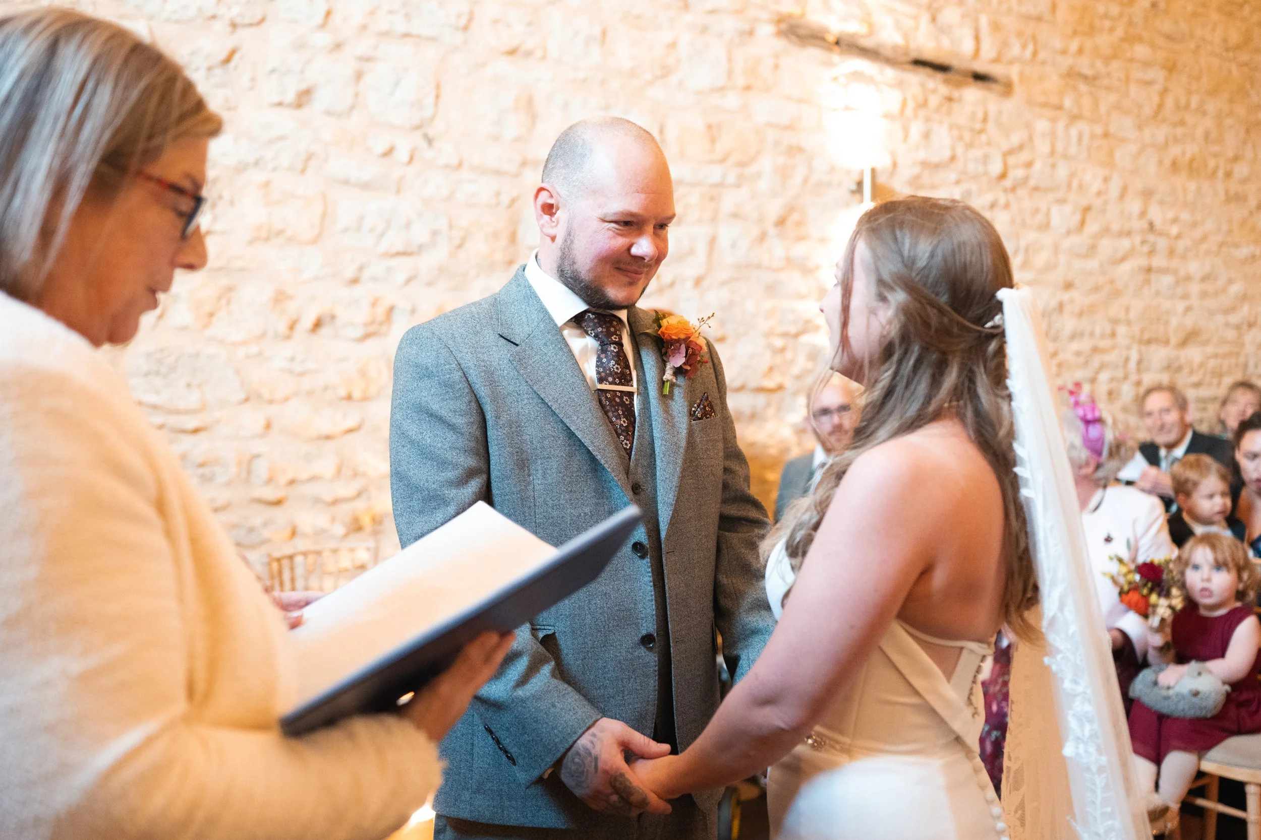 A couple getting married during a wedding ceremony, with a woman officiant reading from a book, in a rustic indoor setting with exposed brick walls.