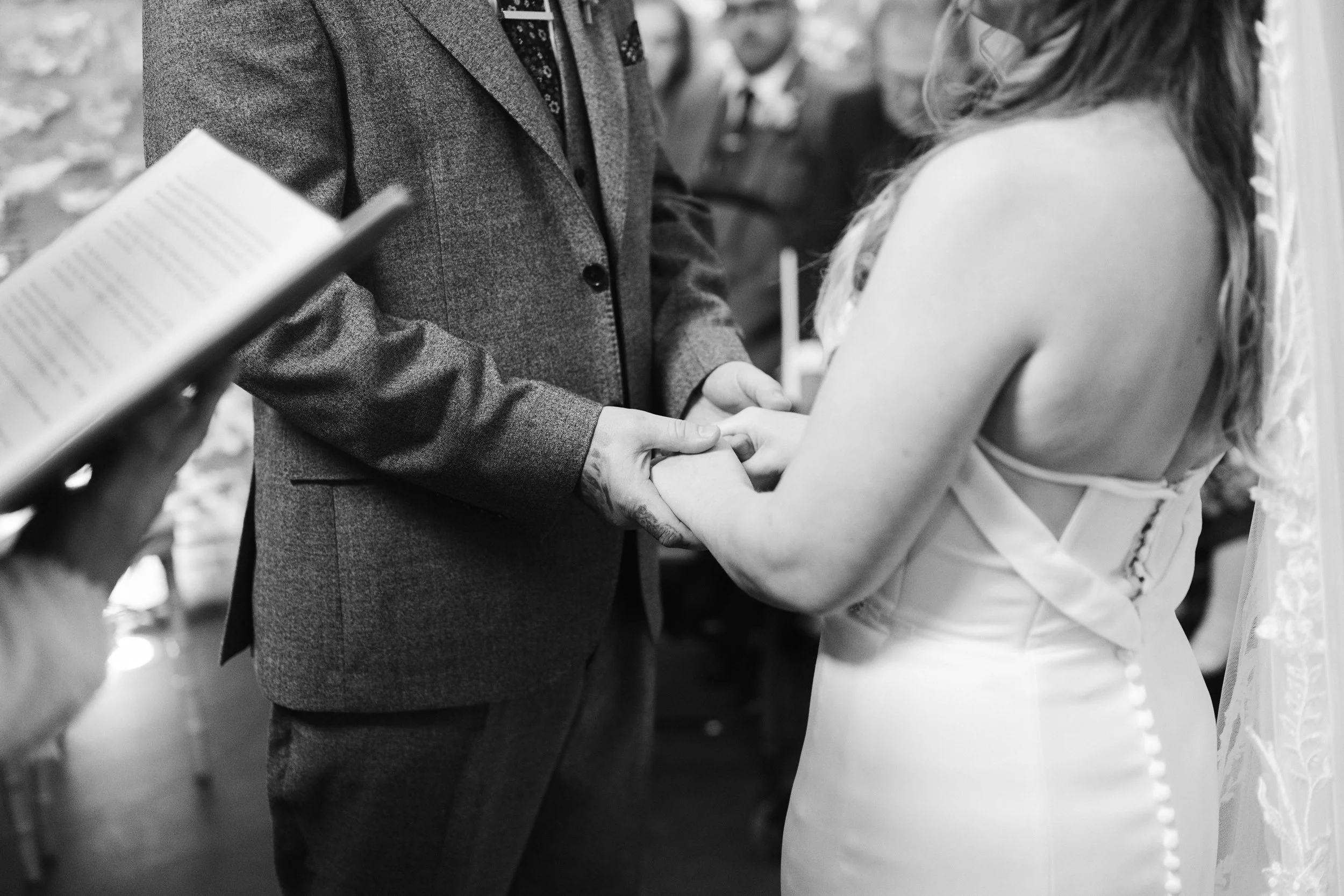 A couple holding hands during a wedding ceremony, with the officiant reading from a book in the foreground. The groom is dressed in a suit and the bride in a white dress, indoors with other people in the background.