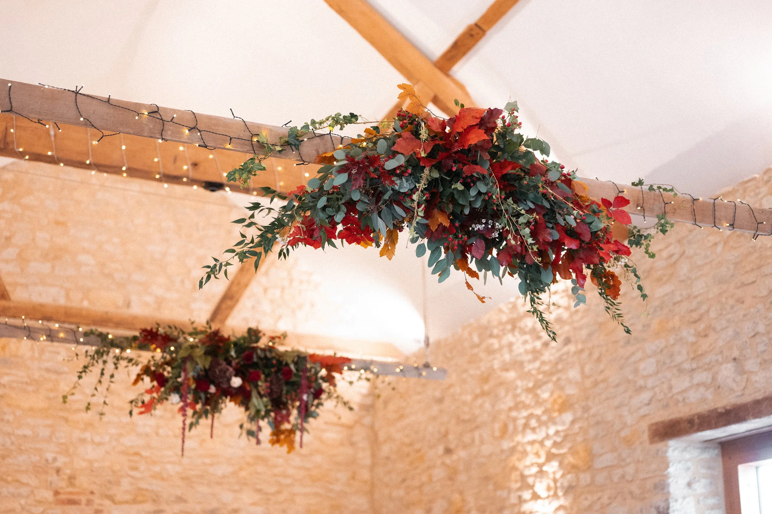 Decorative floral arrangements hanging from wooden beams with string lights in a rustic indoor setting.