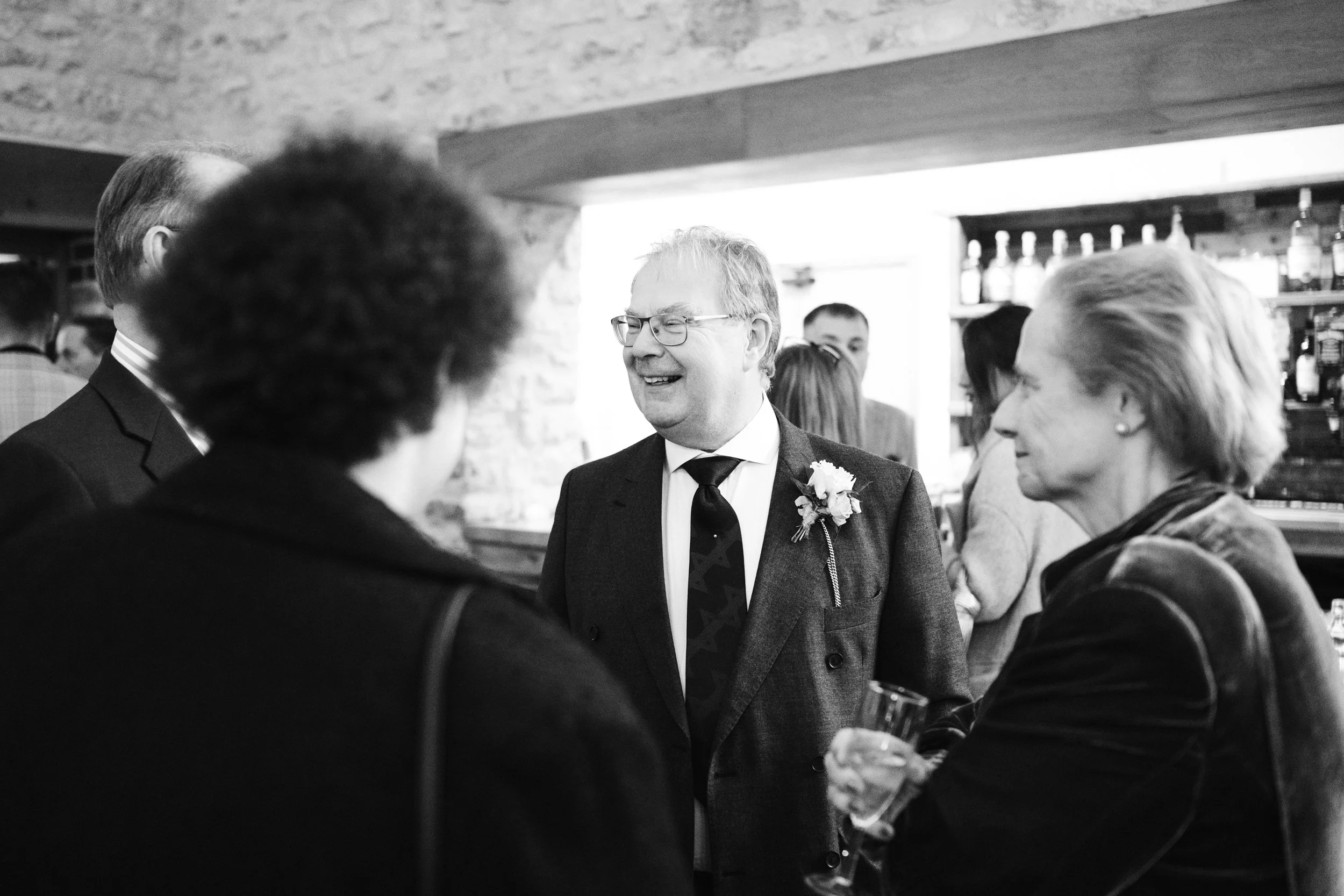 People socializing at a gathering or party in a bar or restaurant, with a man wearing glasses and a suit smiling and engaging in conversation.