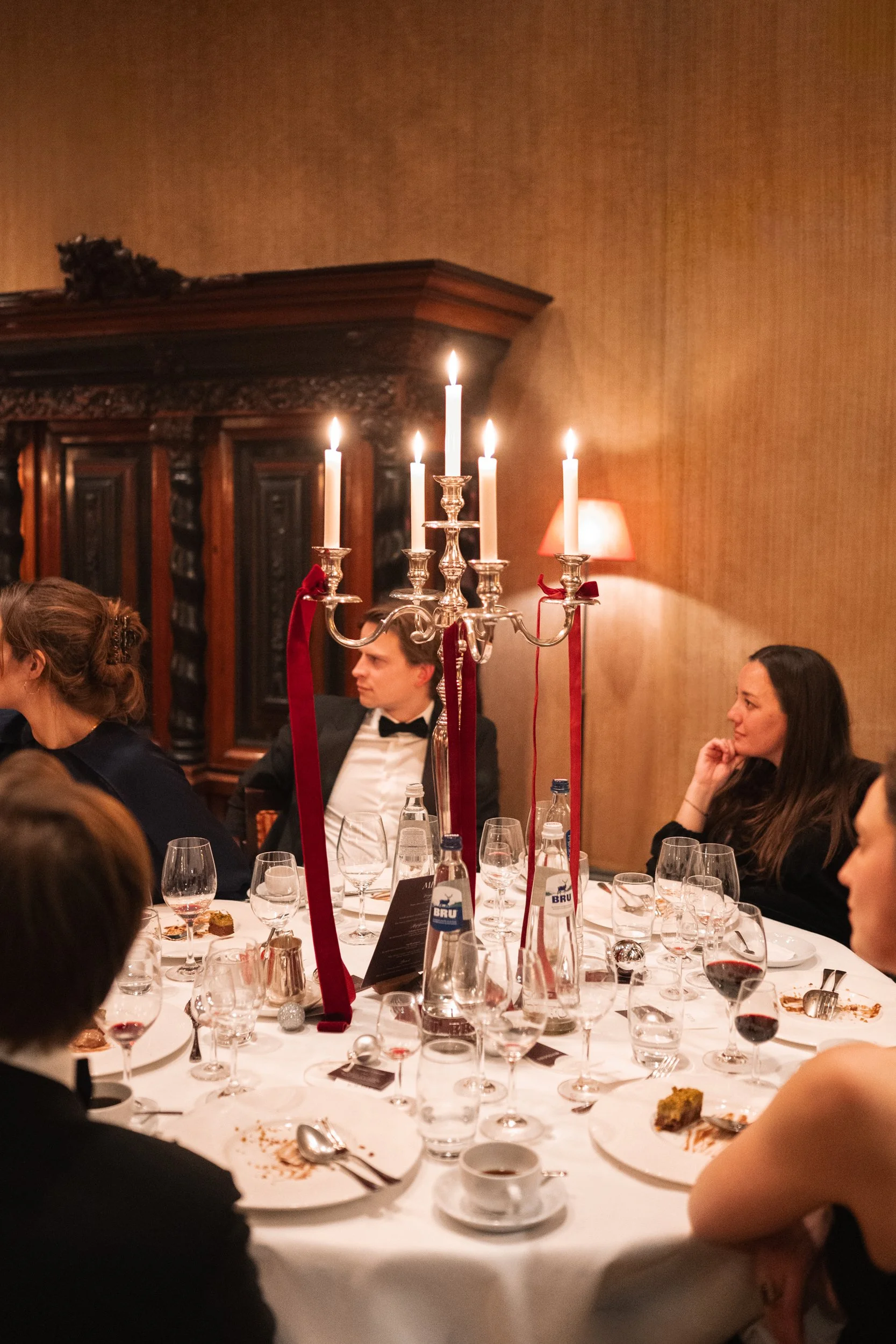 People dining at a formal dinner table with a candelabrum centerpiece, wine glasses, and plates, in an elegant, wood-paneled room.