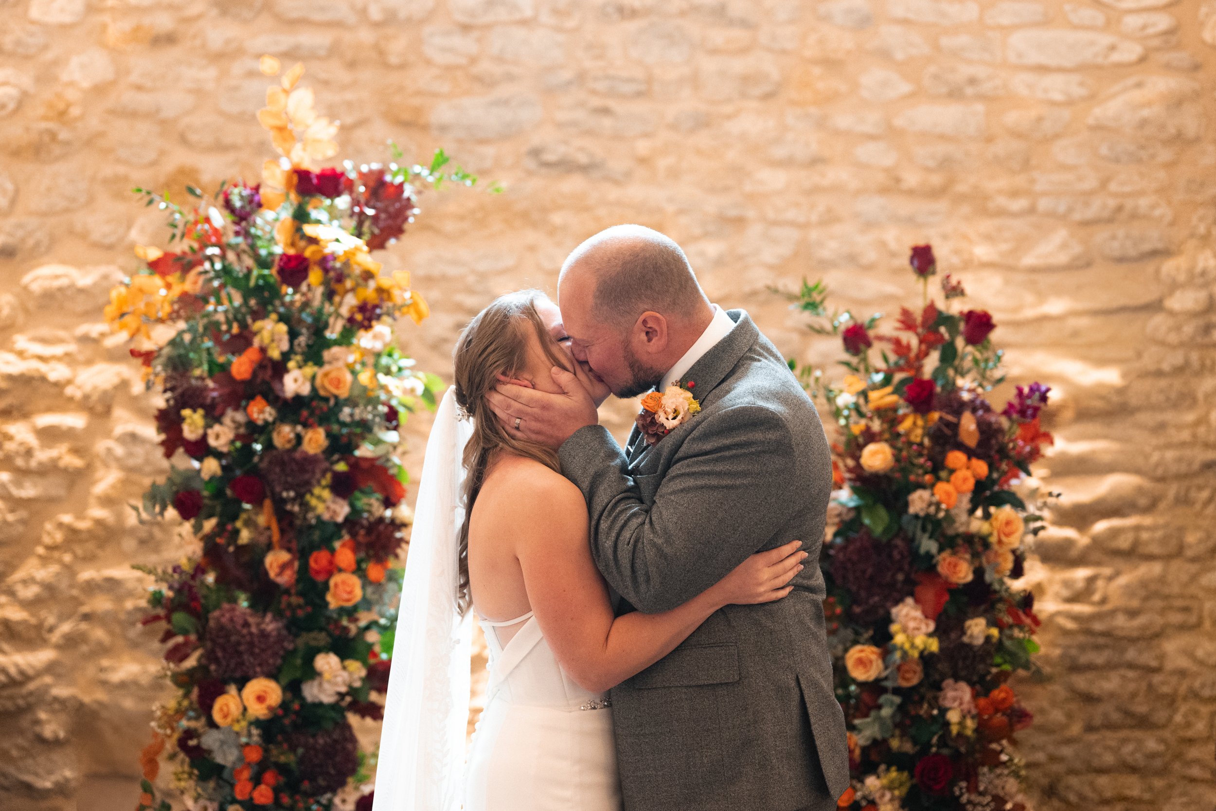 Bride and groom sharing a kiss at their wedding, surrounded by colorful floral arrangements in front of a stone wall.