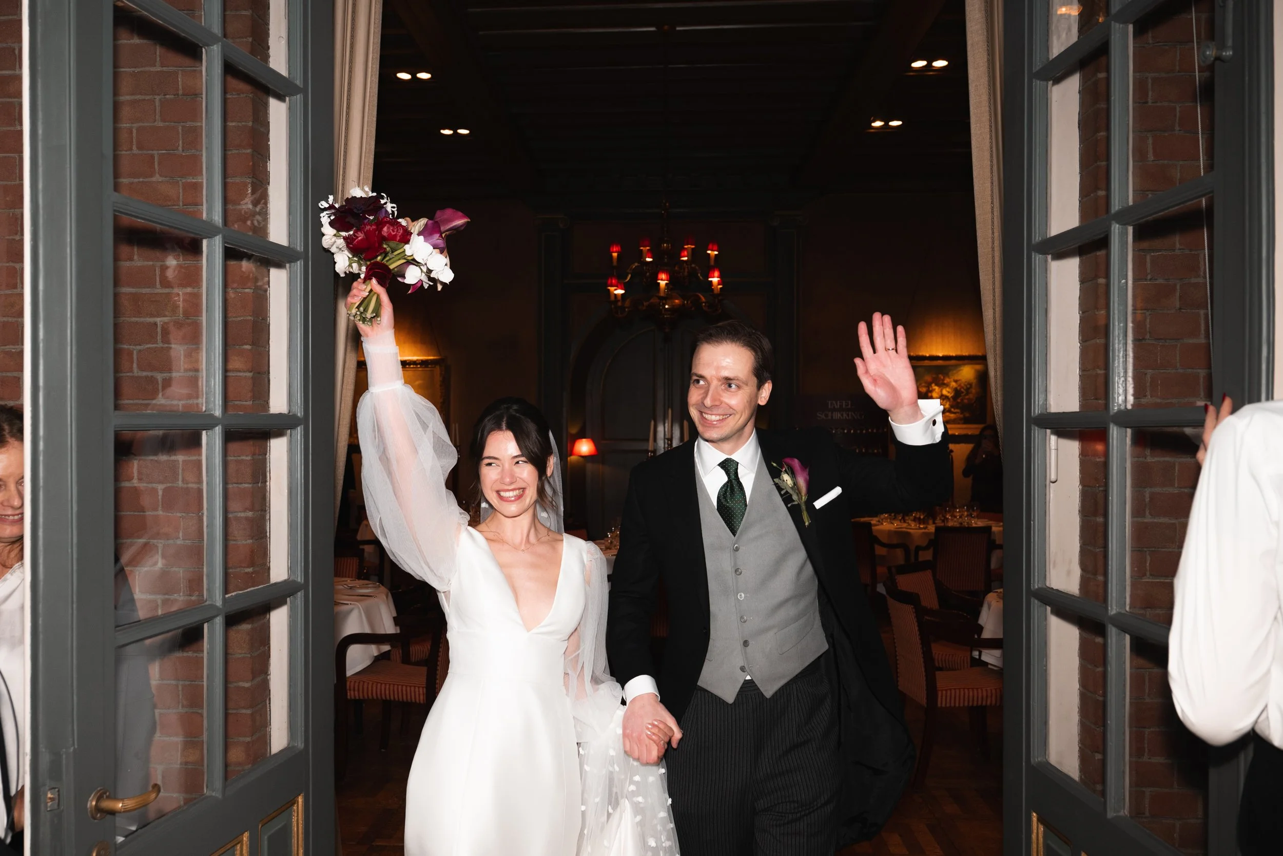 A newly married couple enters a reception hall, smiling and holding hands. The bride is raising her bouquet in the air, wearing a white wedding dress and veil. The groom is waving with his other hand, dressed in a black suit with a grey vest and gree