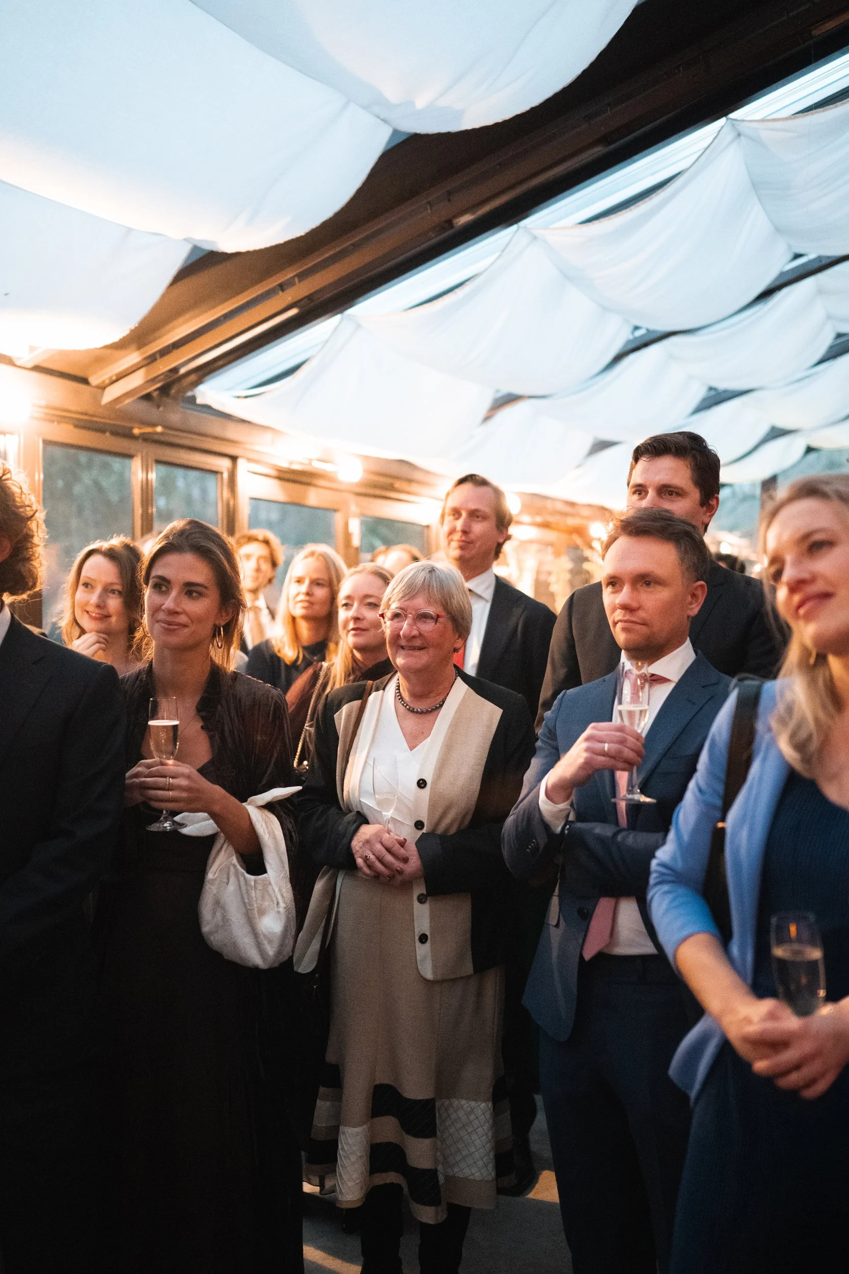 People in formal attire attending an indoor event, some holding glasses of champagne, under a ceiling decorated with white fabric, during sunset or early evening.