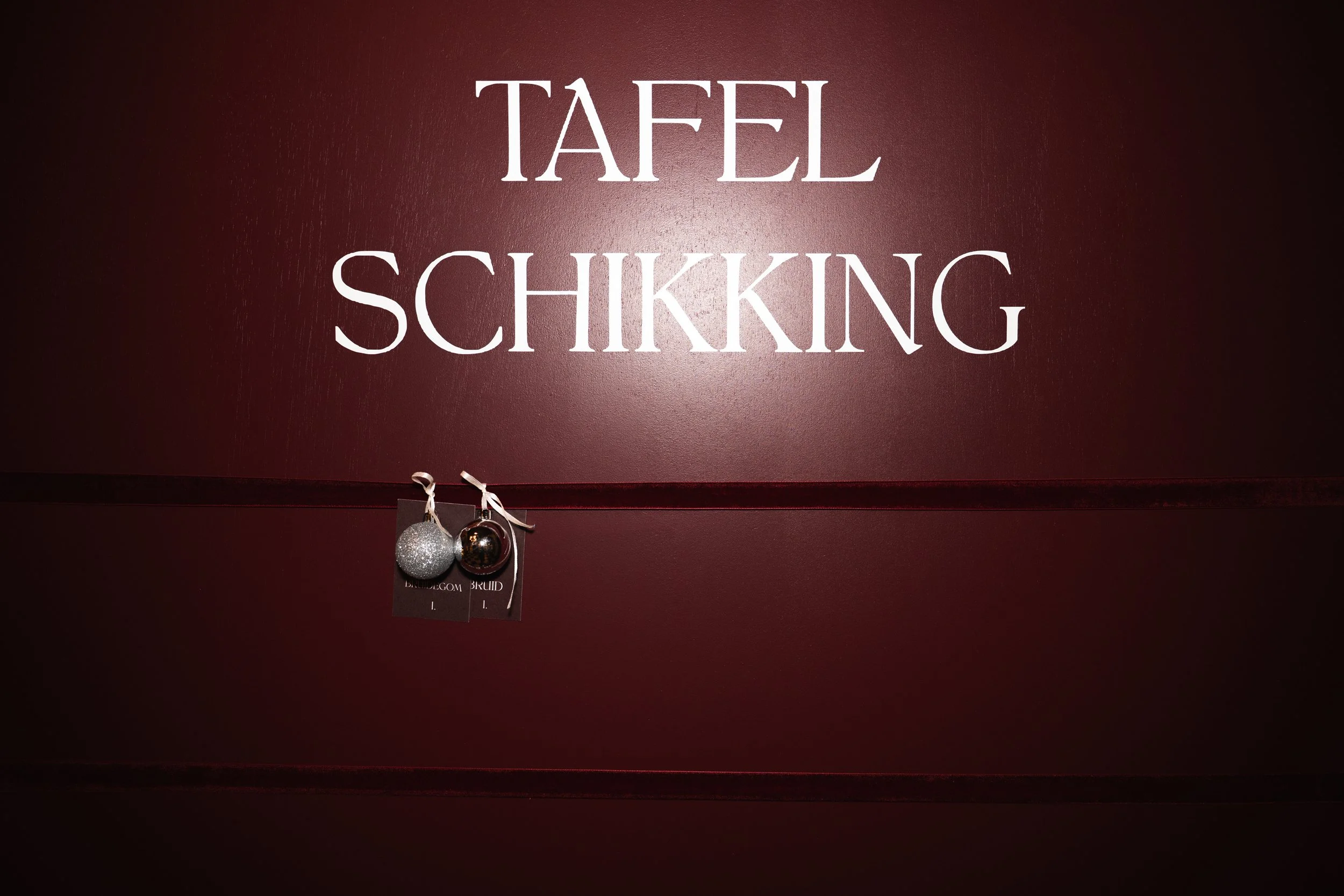 Close-up of a burgundy wall with the words 'TAFEL SCHIKKING' in large white letters and two Christmas ornaments hanging from a string below the text.