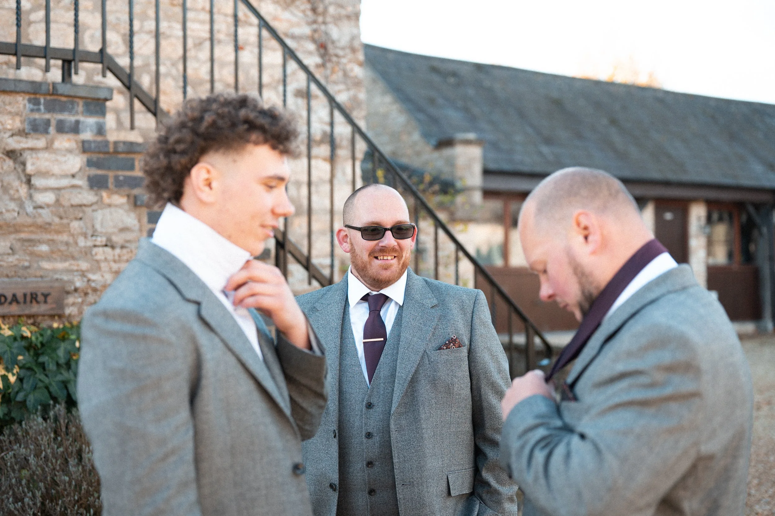 Three men in gray suits stand outside near a brick building, two adjusting their ties and one smiling at them.