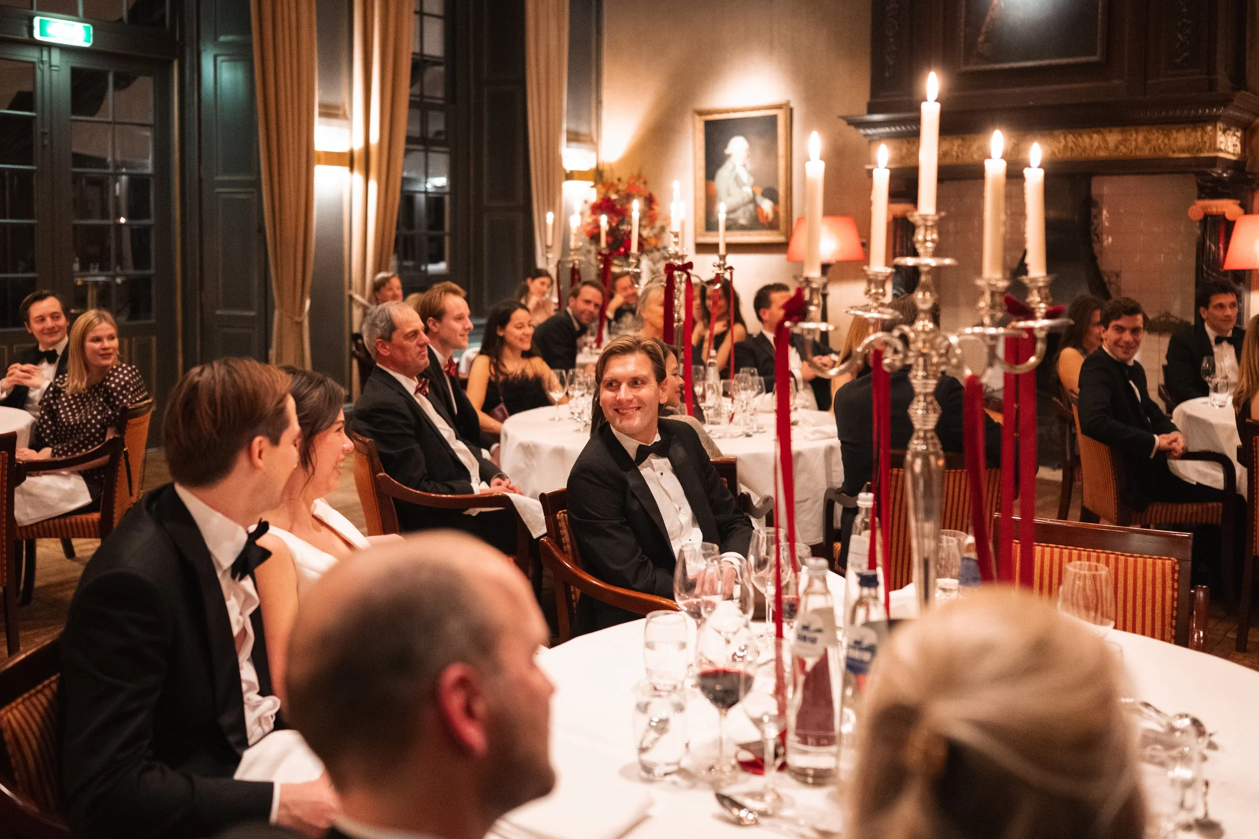 Guests in formal attire attending a dinner event in a decorated ballroom with candles, portraits, and warm lighting.