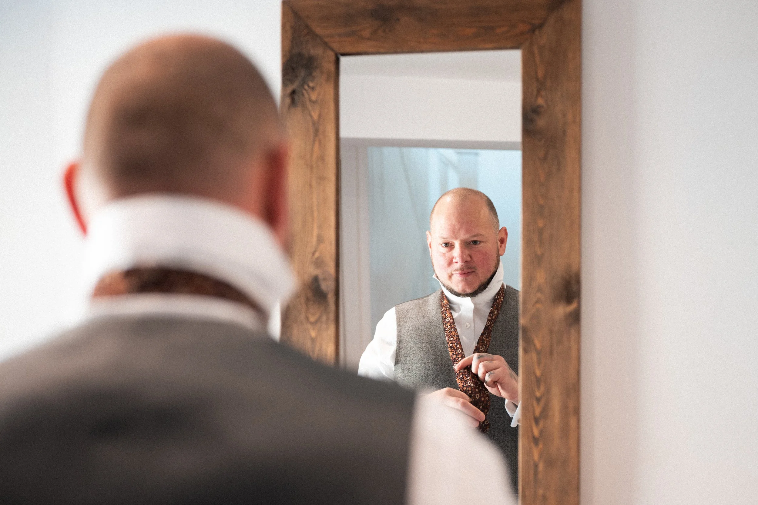 A man is looking at himself in a mirror while adjusting his tie, wearing a gray vest and white shirt.