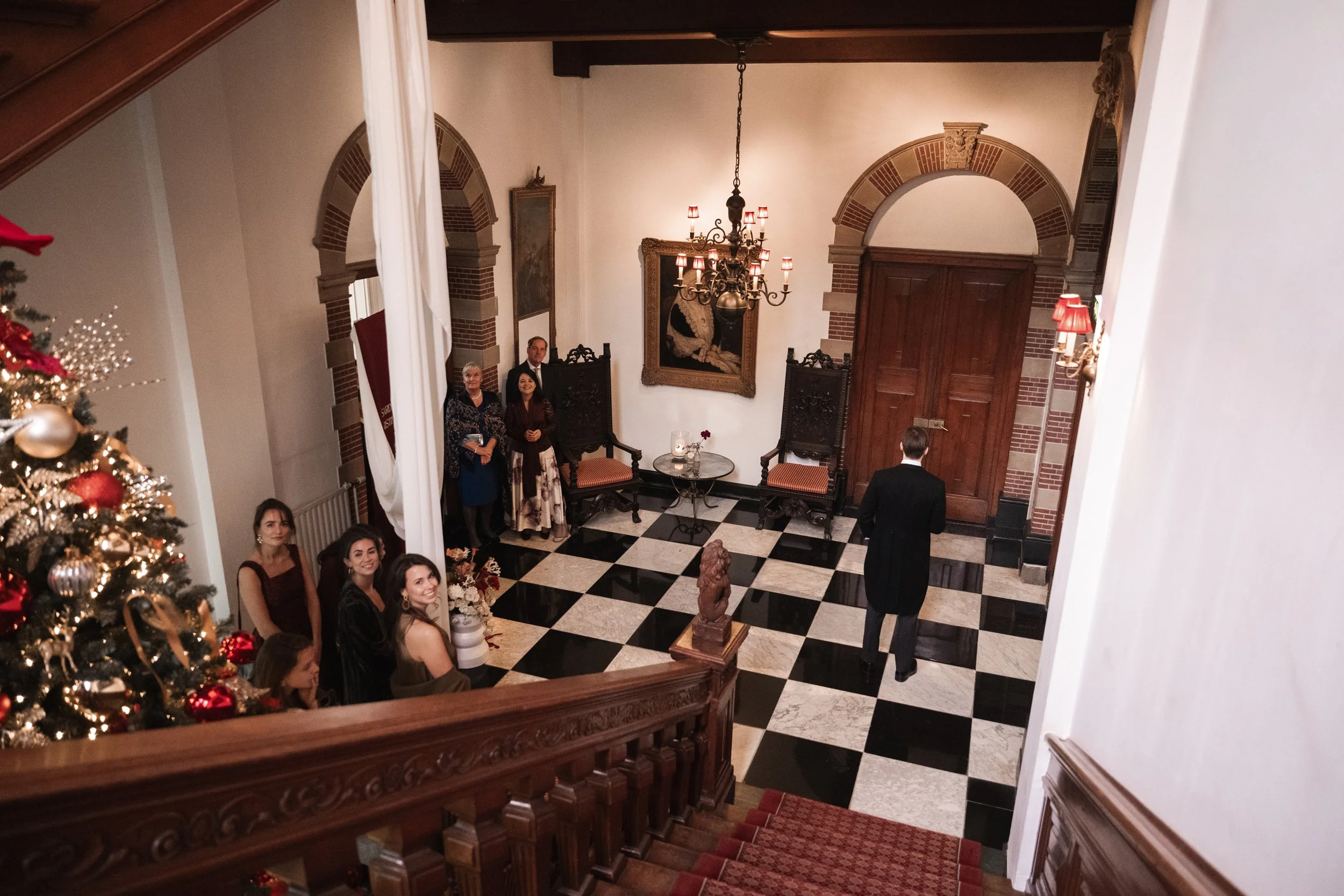 People gathered in an elegant foyer with a black and white checkered floor, a large chandelier, and wooden accents, during a formal event or celebration.