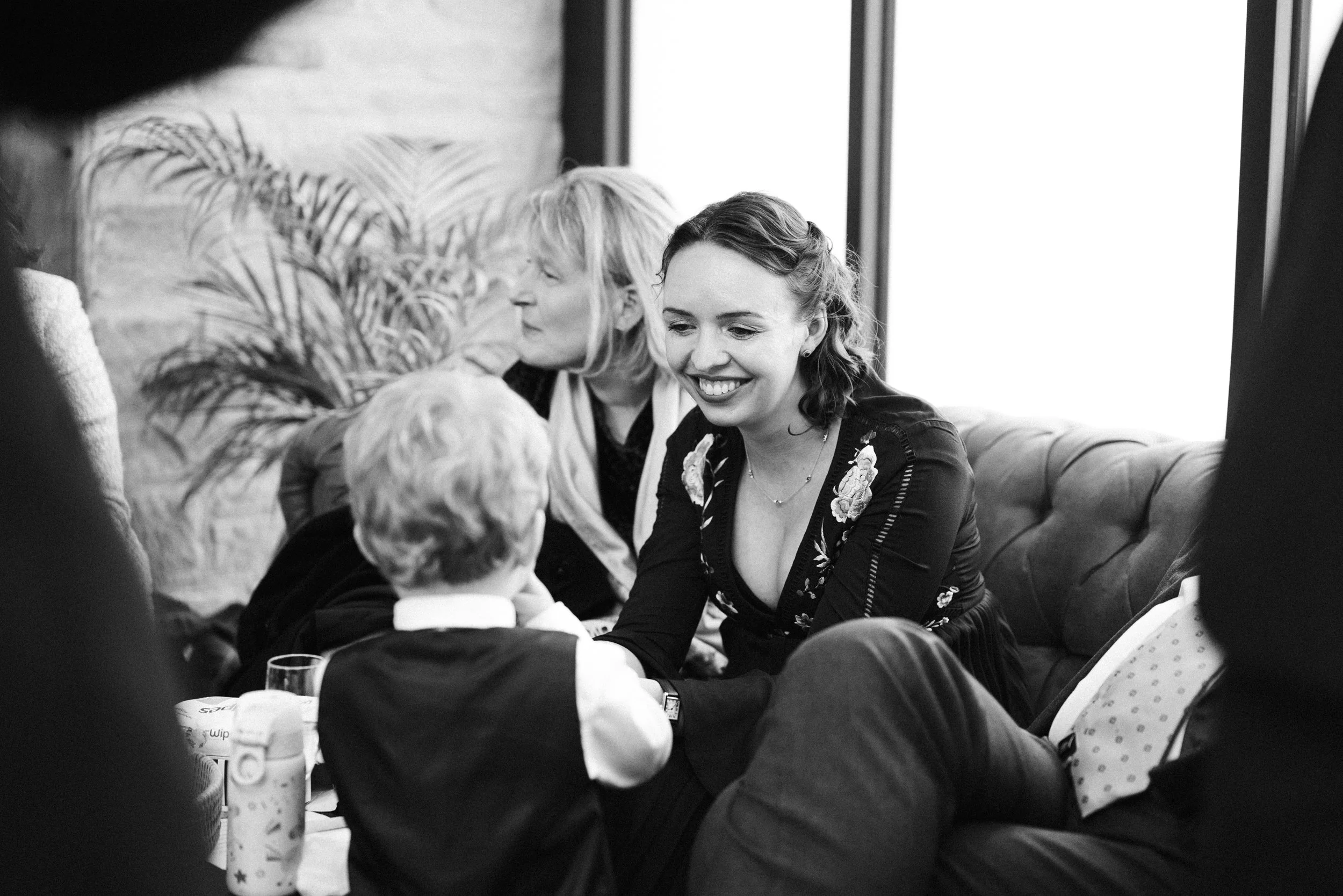 A woman smiling and talking to a young boy in a black vest, in a social setting. Other people are in the background, with a woman and a large plant visible, sitting on a sofa near a window.