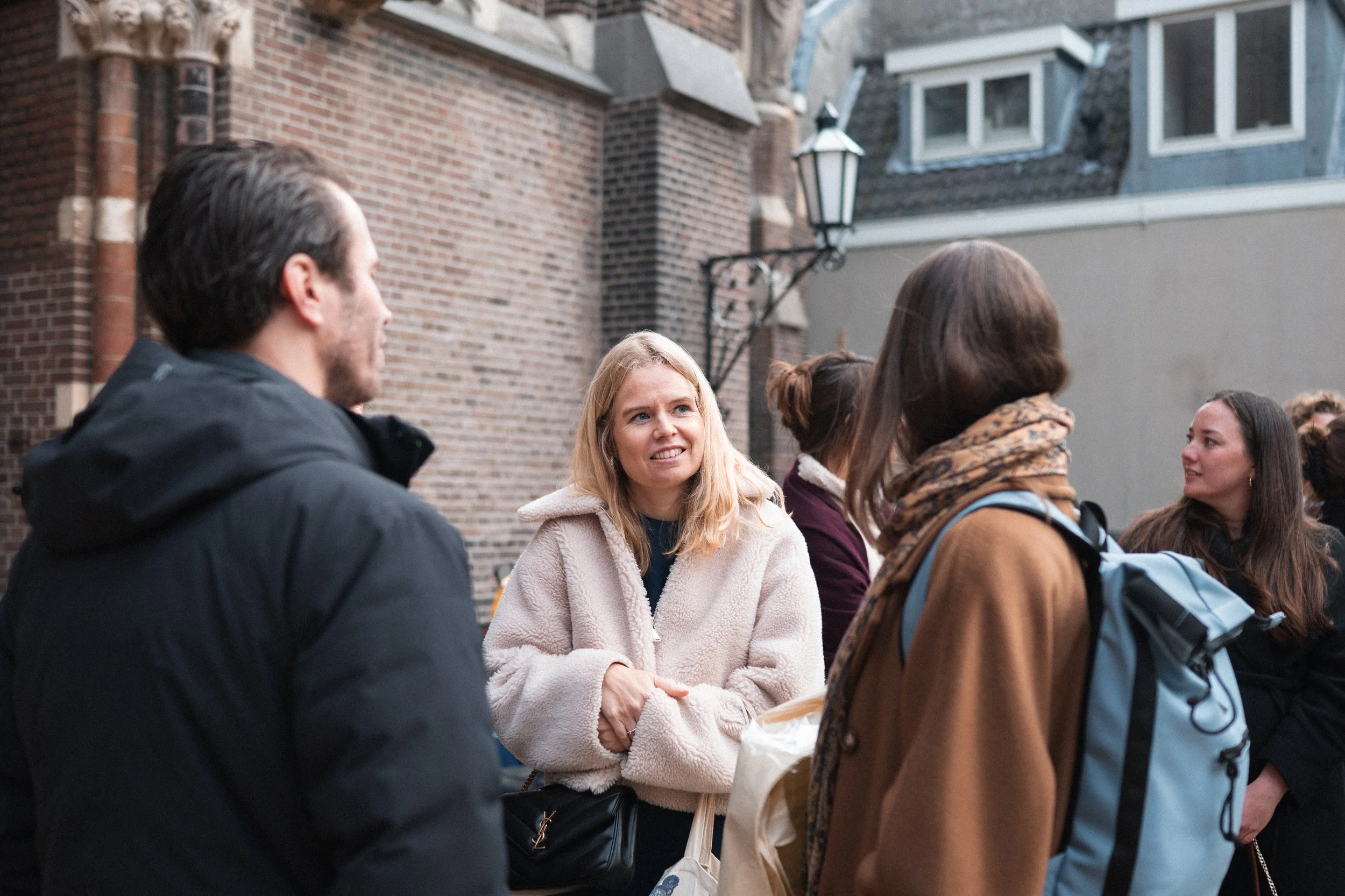 Group of people talking outside near brick building, some wearing coats and backpacks, in daylight.