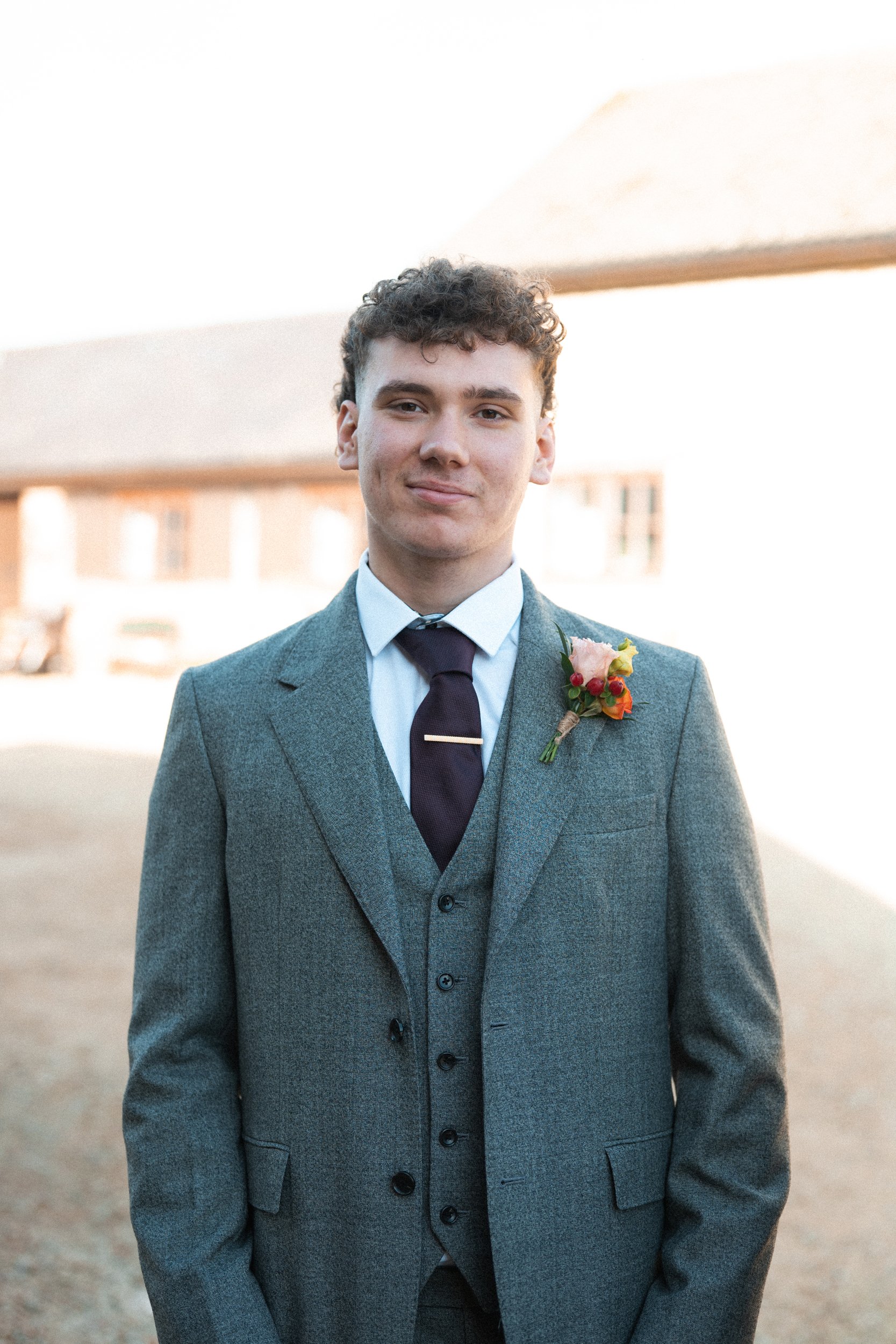 A young man in a gray suit with a boutonniere, white shirt, and dark tie, standing outdoors with sunlight, smiling at the camera.