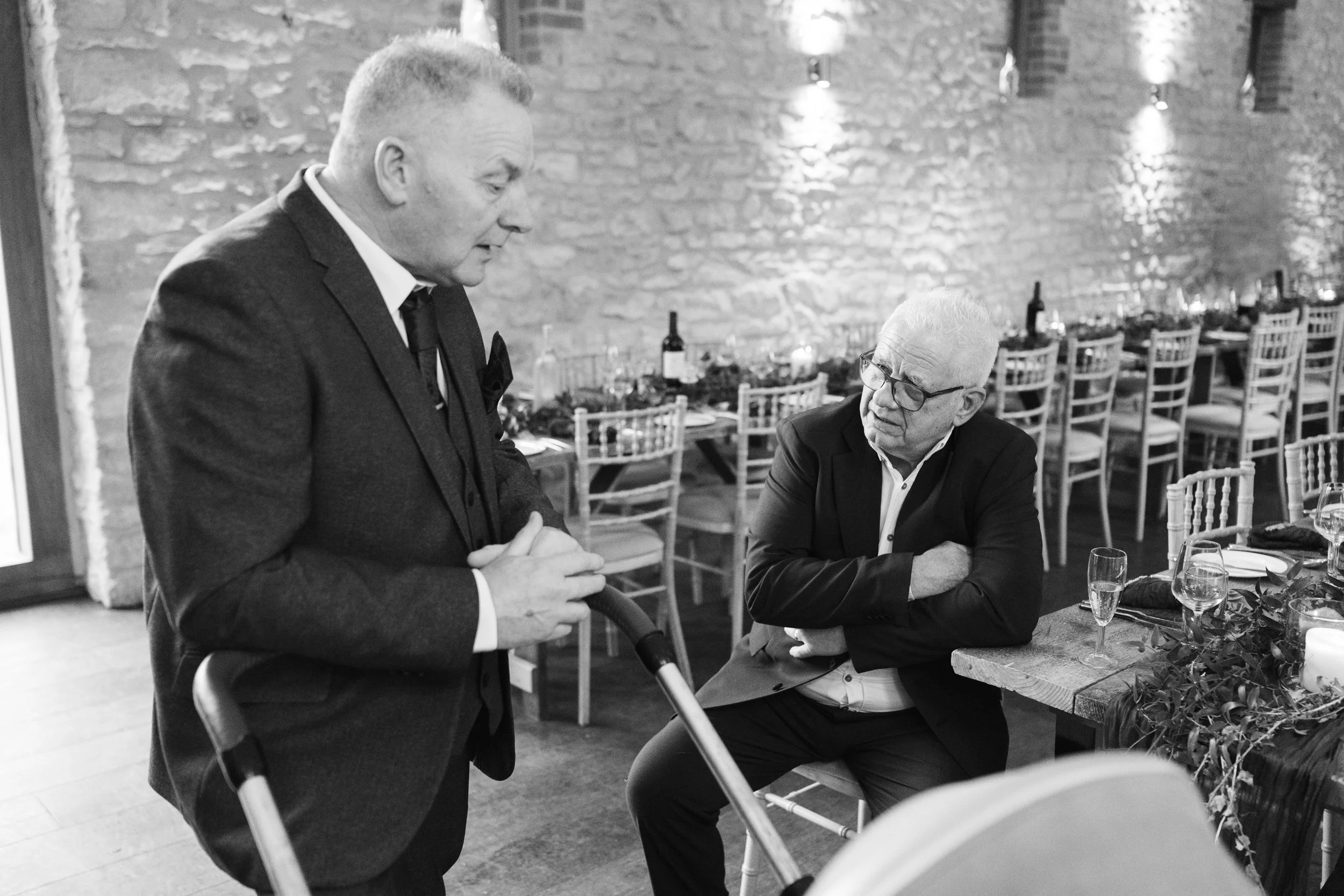 Two men in suits having a conversation in a restaurant with brick walls and decorated tables.