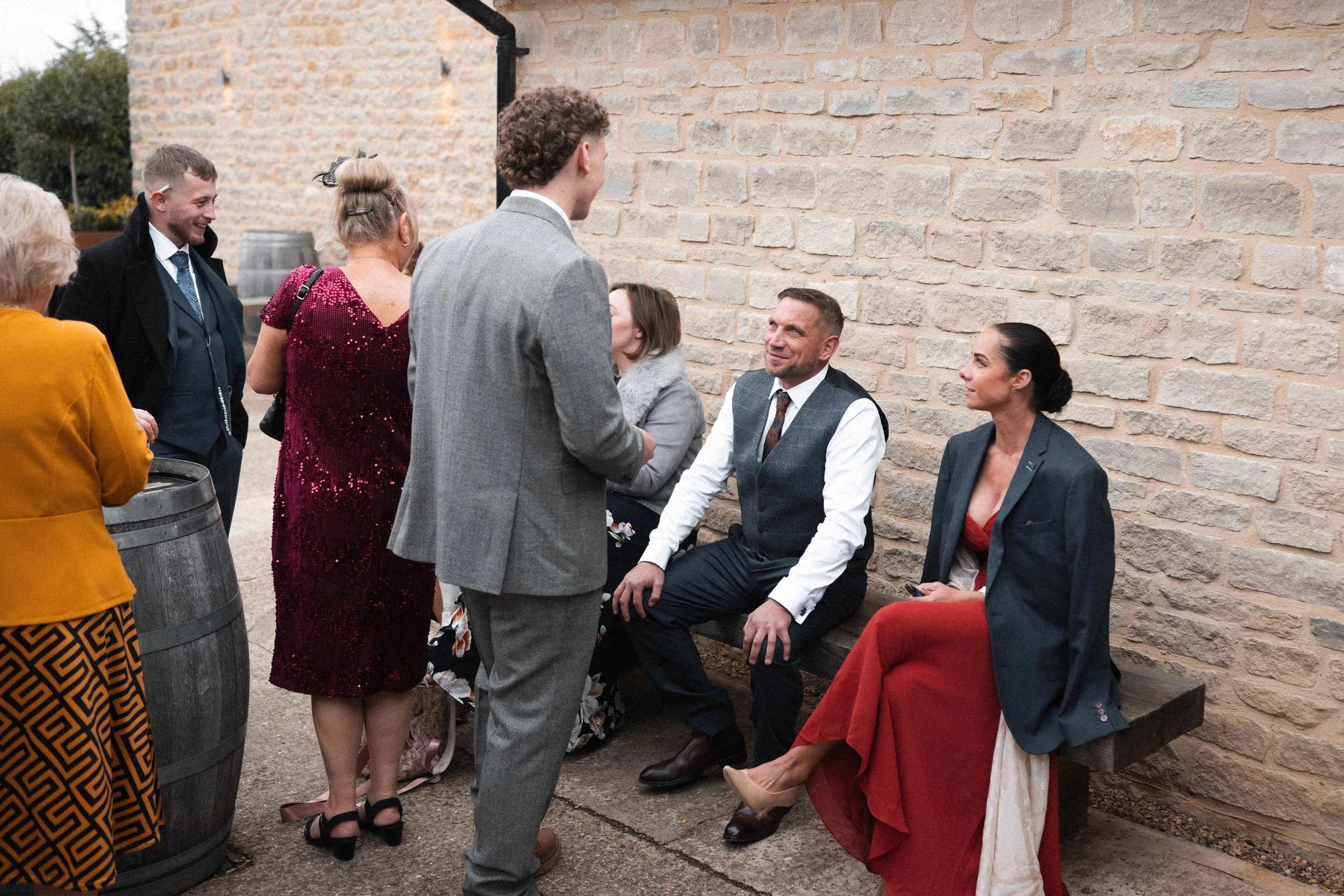 People gathered outside near a brick wall and wine barrels, engaged in conversation. Some are seated, others standing, dressed in formal attire.
