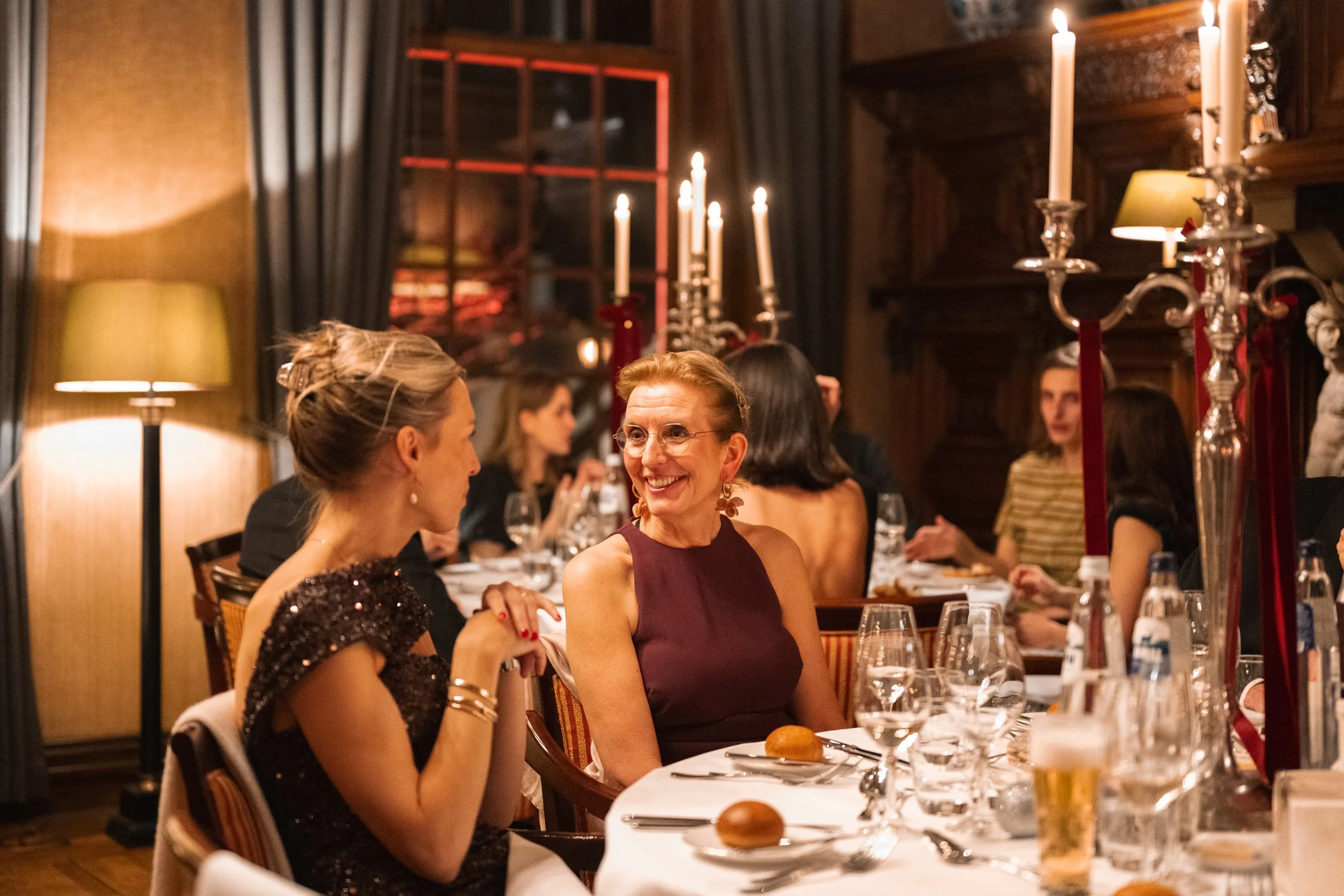 A woman in a dark purple sleeveless dress smiling and talking to another woman with a bun hairstyle, wearing a dark, possibly sequin dress, at a formal dinner party with multiple tables, wine glasses, candles, and ornate decor.