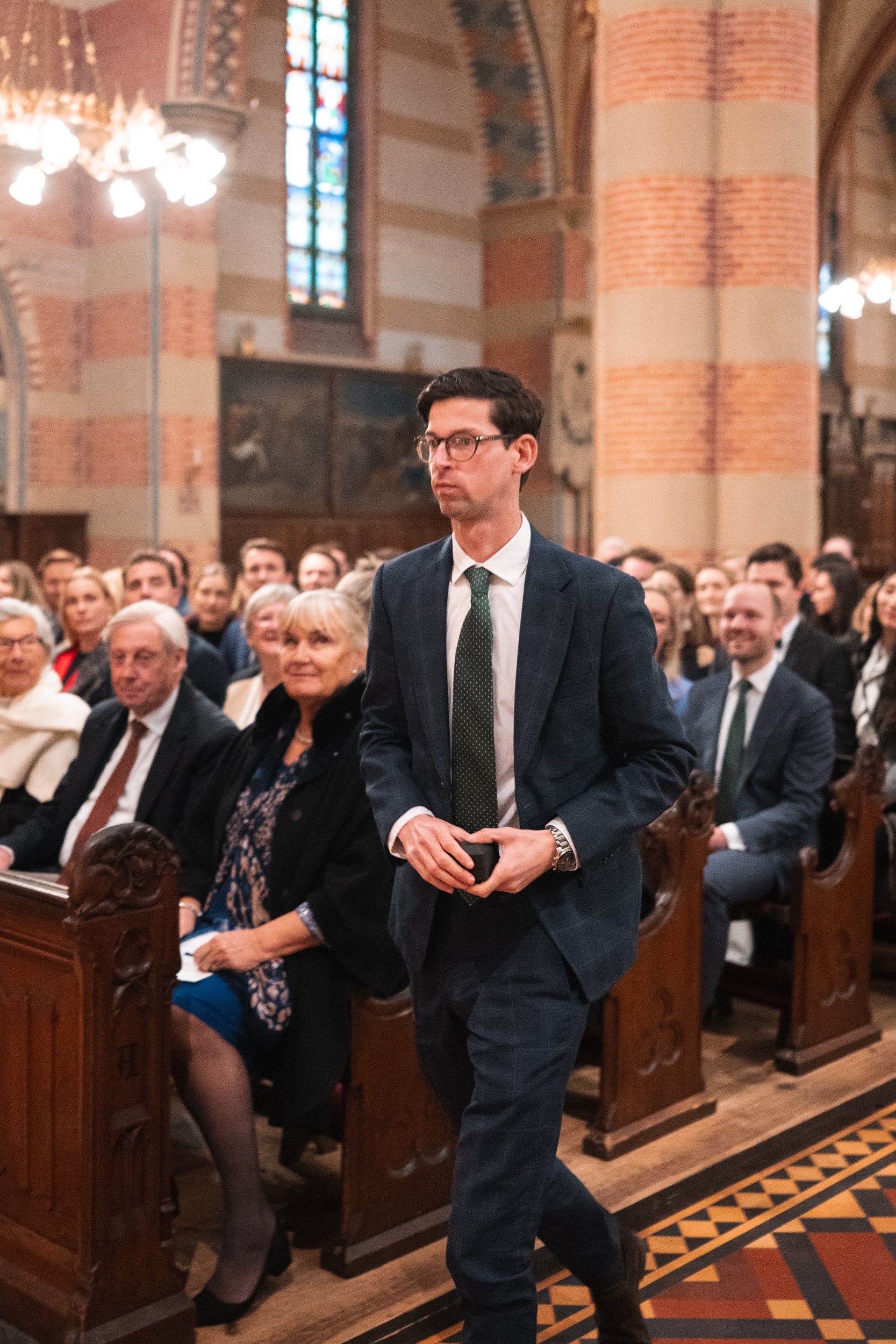 A man in a dark suit, white shirt, and glasses walking in a church filled with seated people, some smiling, with stained glass windows and ornate architectural details in the background.