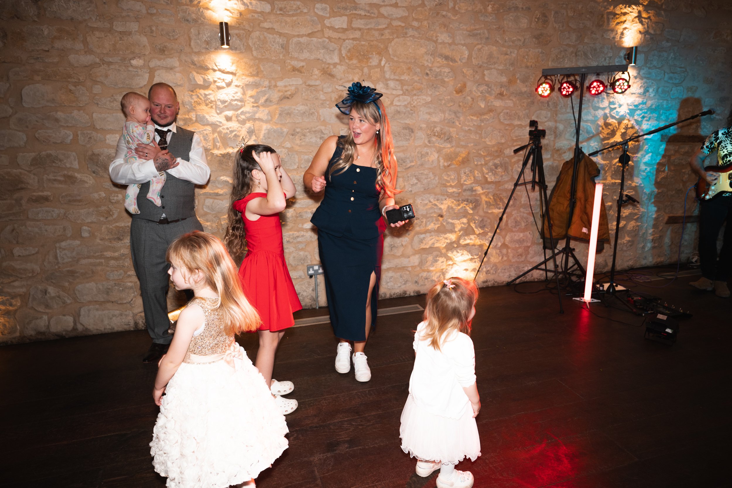 A woman in a navy dress and hat dancing at a party with children in fancy dresses on a dark wood floor against a stone wall background.