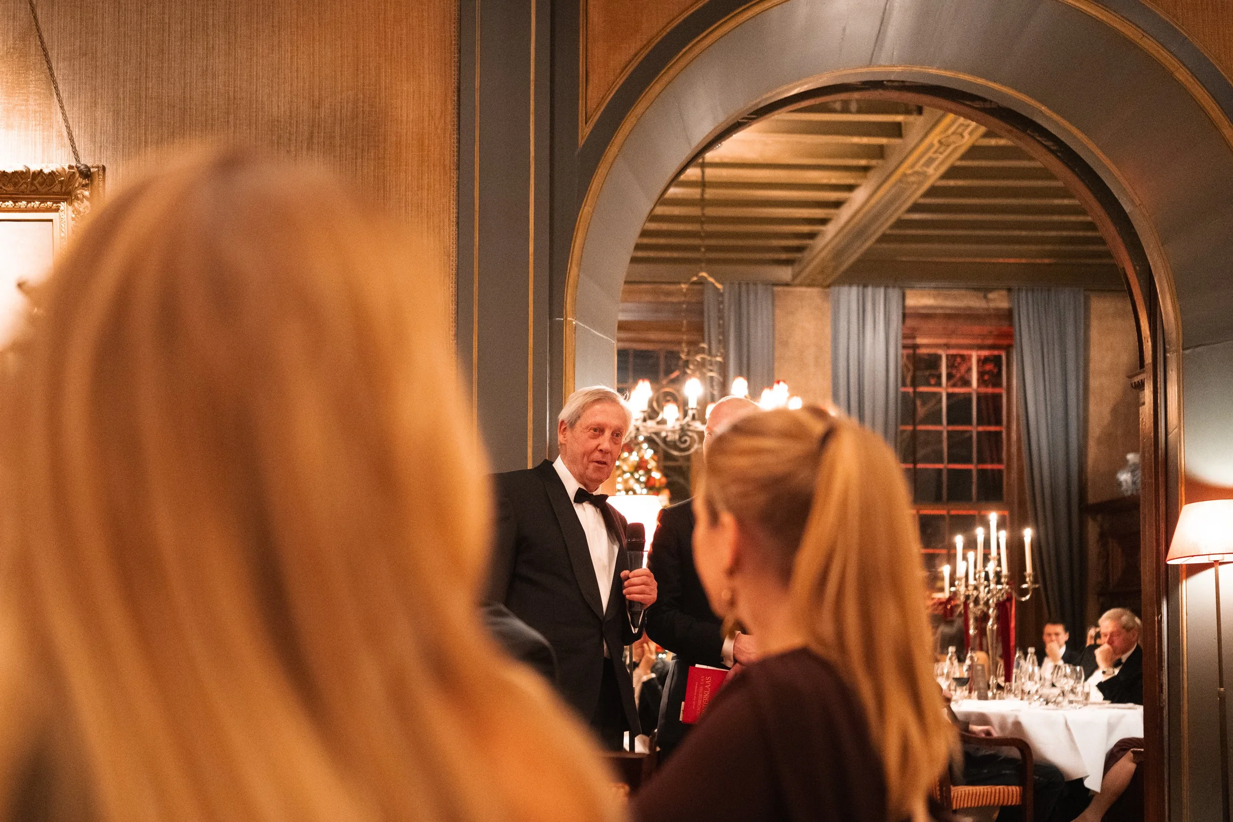 An older man in a tuxedo speaking into a microphone at a formal dinner event, with guests seated at tables decorated for Christmas in the background.