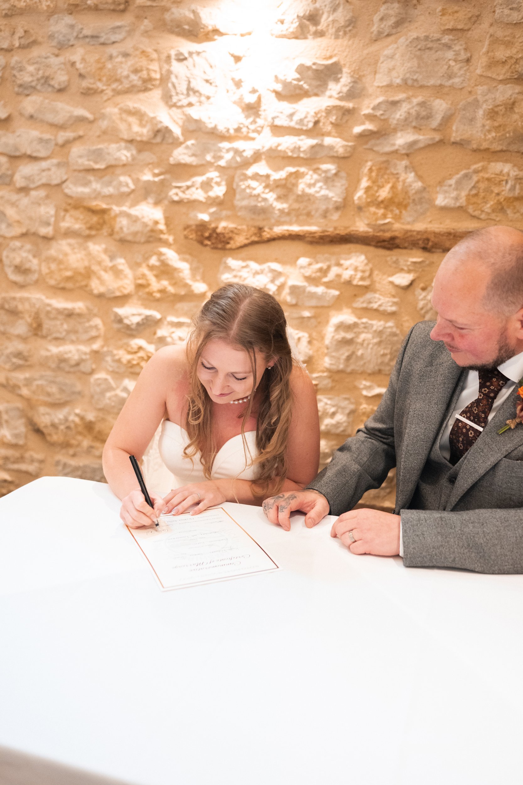 A bride signing a marriage license with her groom sitting beside her during a wedding ceremony in a rustic venue.