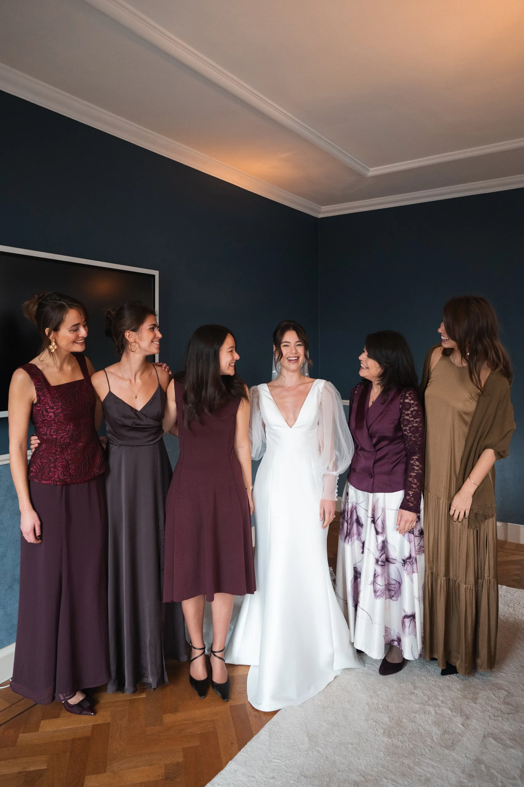 Group of women standing together indoors, dressed in formal attire, with the bride in a white wedding gown at the center, smiling and sharing a joyful moment.