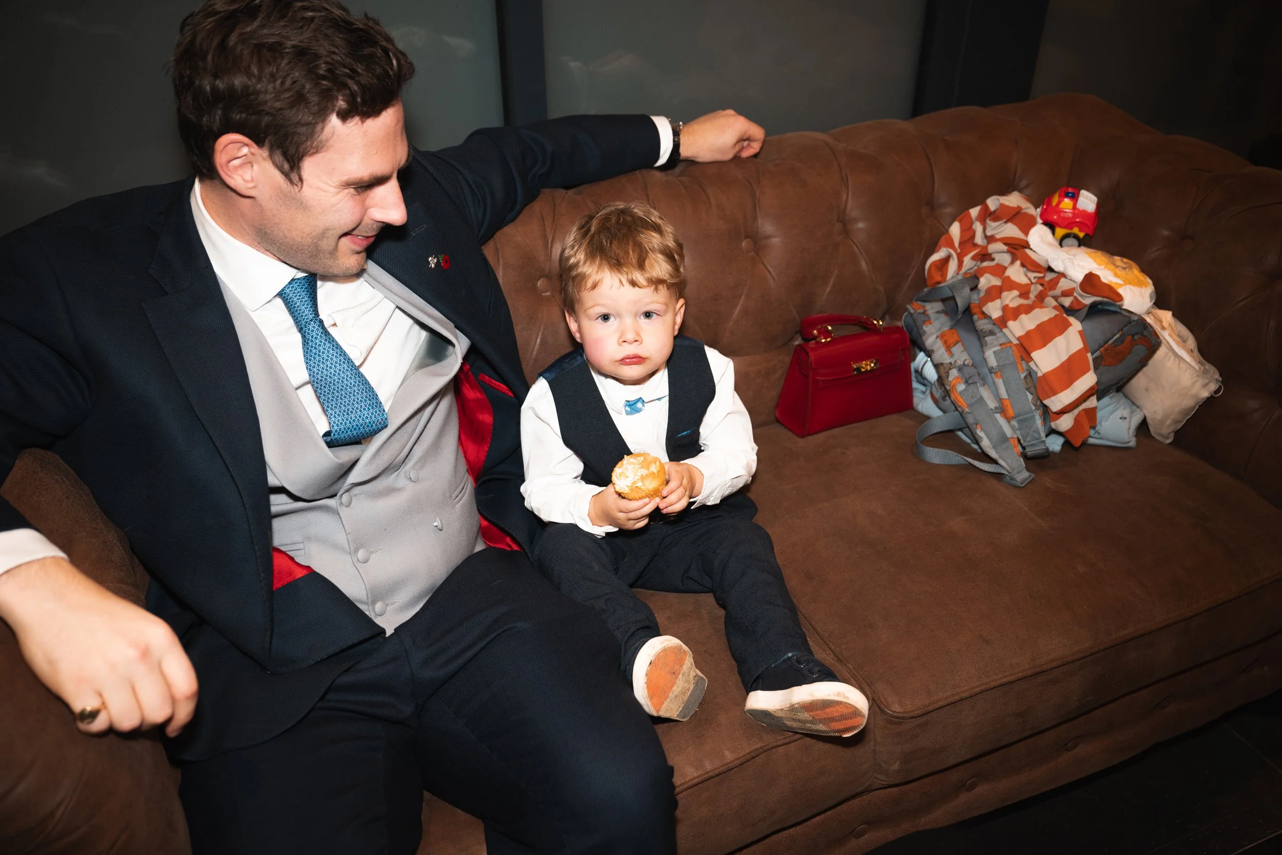 A man in a suit and young boy with blond hair sitting on a brown leather couch. The boy holds a donut and looks at the camera. There are bags and toys on the couch next to them.