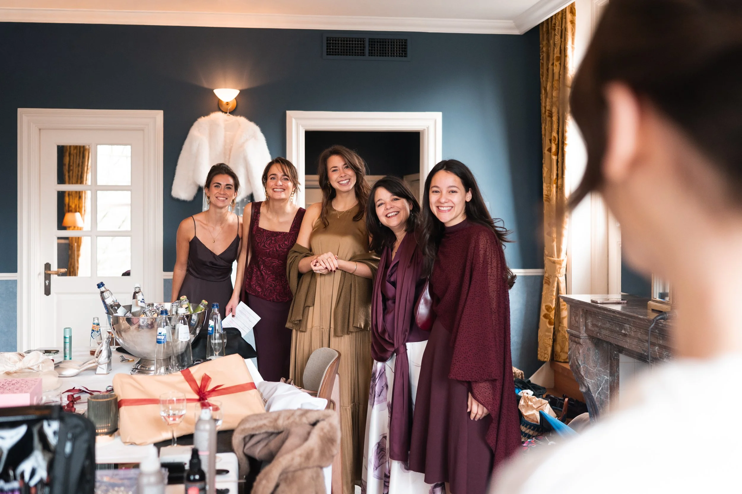 Five women dressed in formal attire smiling and posing for a photo in a room with blue walls, gold curtains, and a white fur coat hanging on the wall.