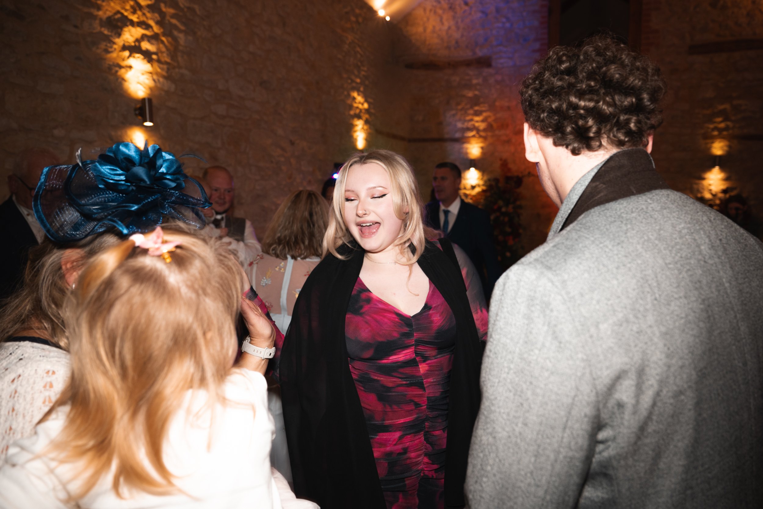 Group of people socializing at a party in a warmly lit room with stone walls, including a woman with blonde hair smiling and talking to a man with curly hair and a gray jacket, while a girl with a large blue hat interacts with them.