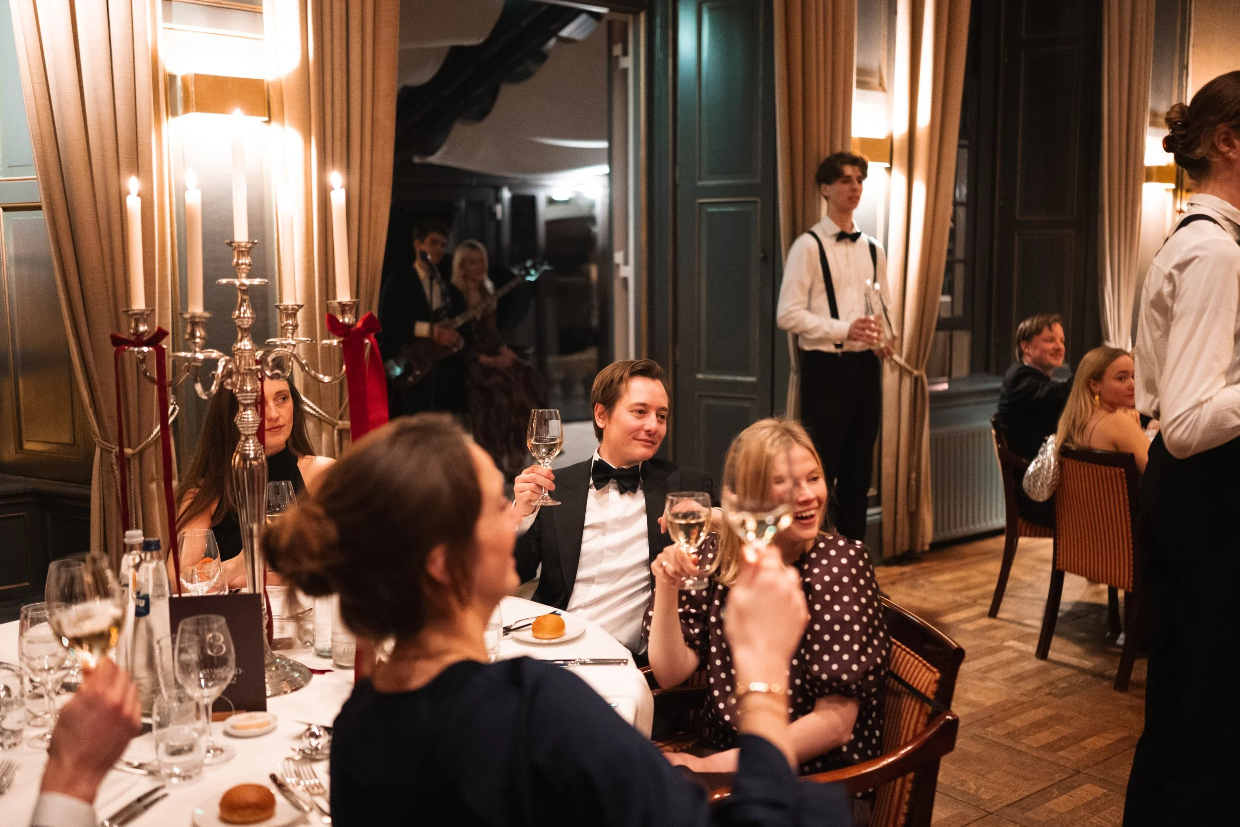 Guests celebrating at a formal dinner party with champagne glasses, elegant attire, and live music in a decorated room with candles.
