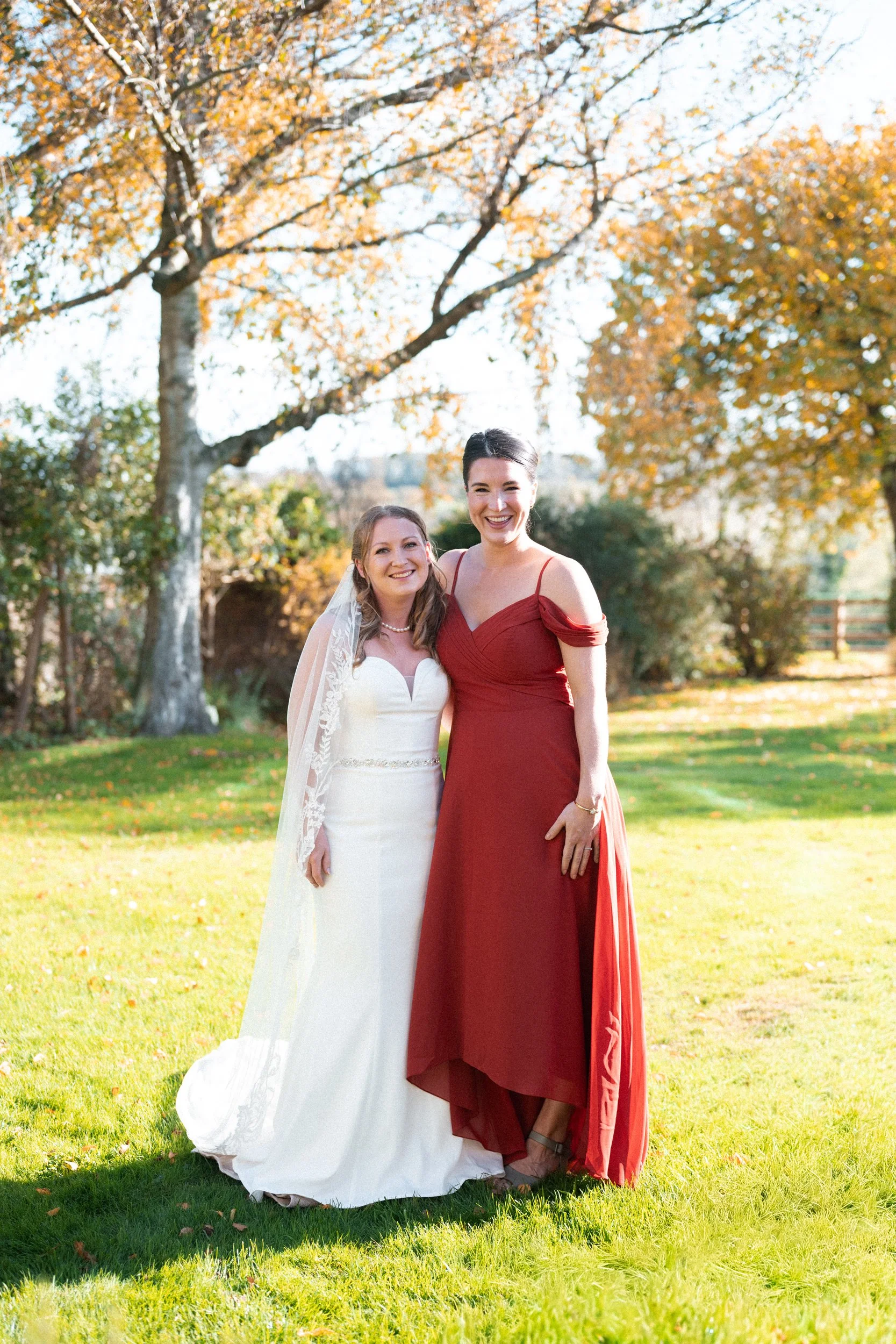 Two women standing together outdoors on a grassy area with trees showing autumn leaves. One is wearing a white wedding dress with a veil, and the other is wearing a red dress. They are smiling.