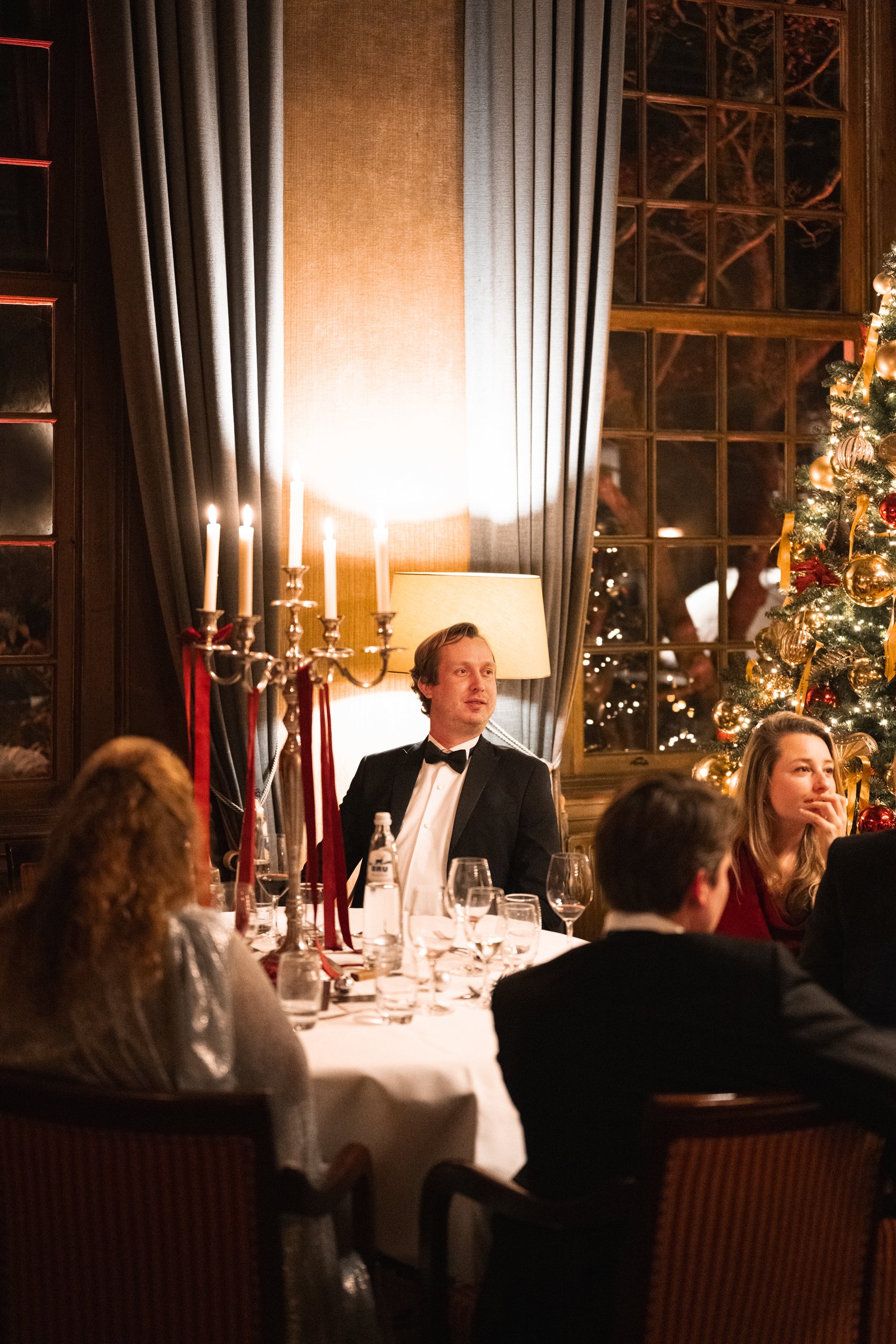 A festive holiday dinner scene with people seated at a round table, decorated with a candelabra, glasses, and a Christmas tree with ornaments, in a warmly lit room with large windows and curtains.