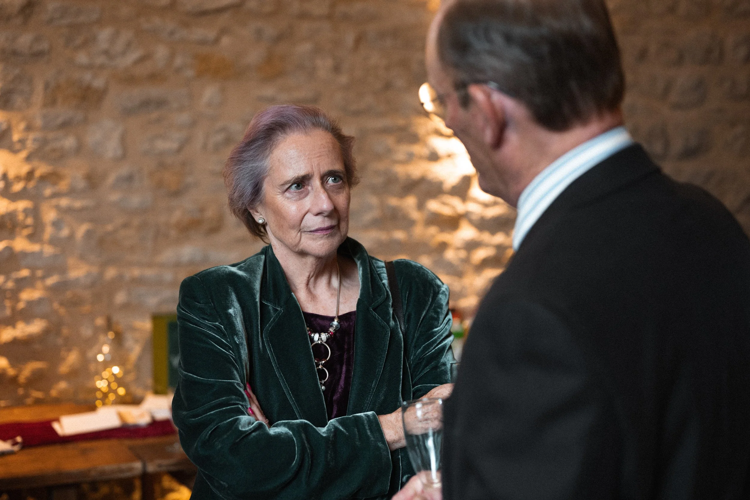An older woman with short gray hair, wearing a green velvet blazer and a necklace, is having a conversation with a man whose face is not visible. The man is dressed in a dark suit and light shirt, holding a glass of champagne. They are indoors with a