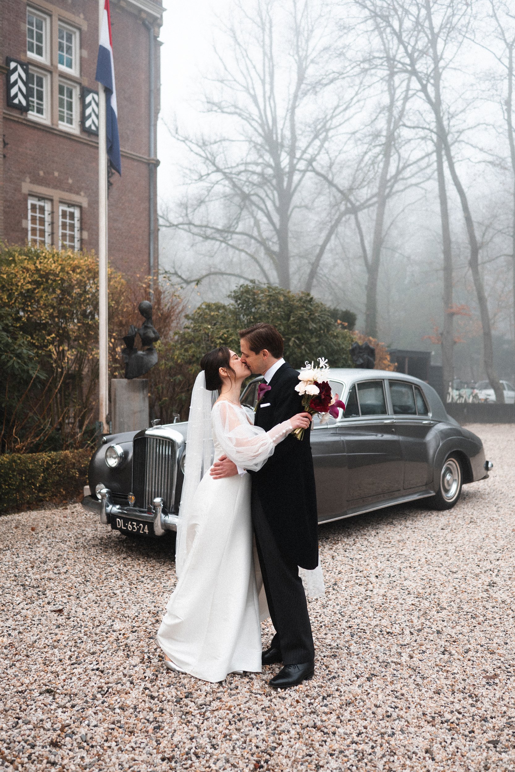 A bride and groom kissing outdoors on their wedding day, with a vintage black limousine and a foggy background.