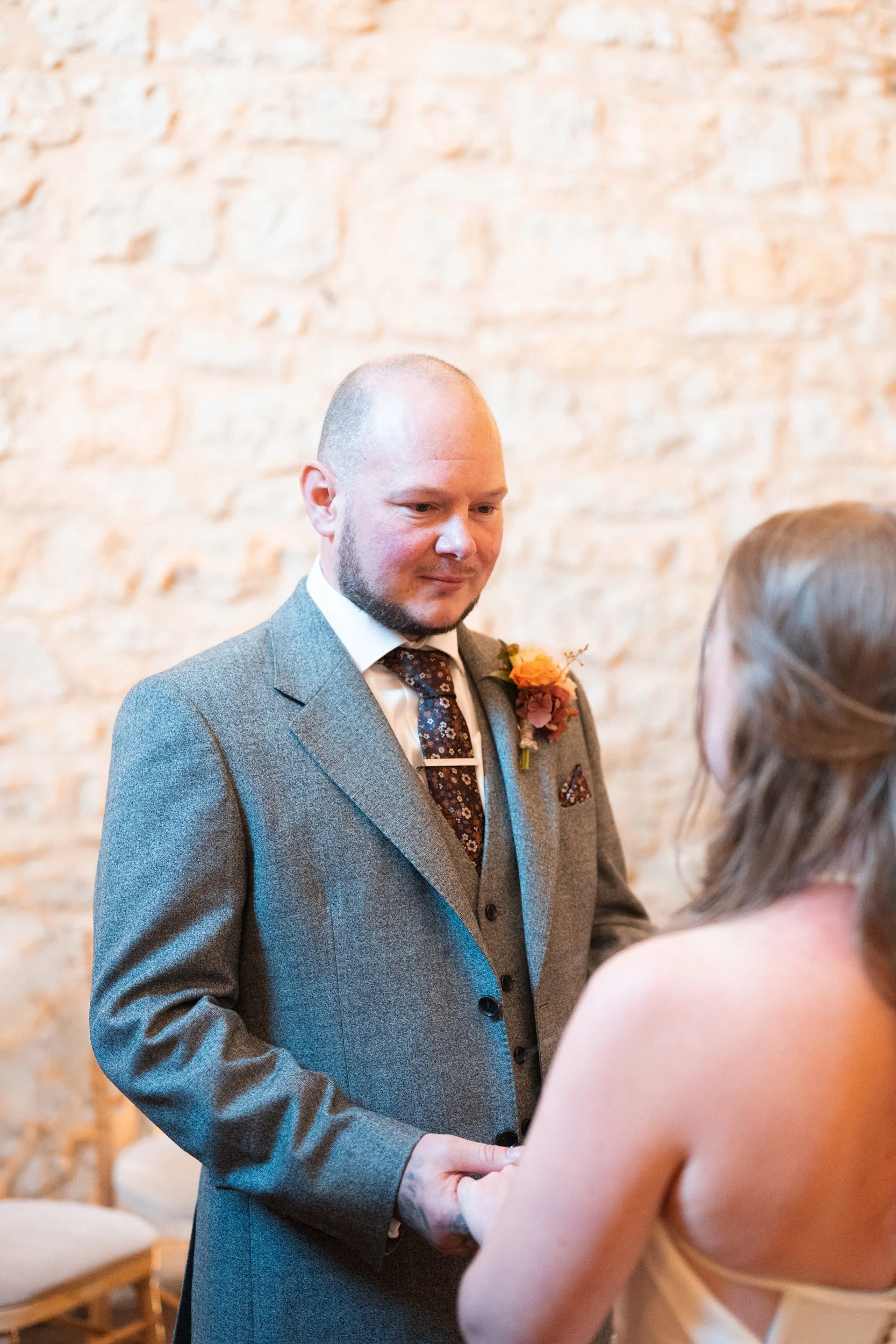 A man in a gray suit with a floral tie and orange boutonniere holding hands with a woman in a cream dress during a wedding ceremony in front of a brick wall.