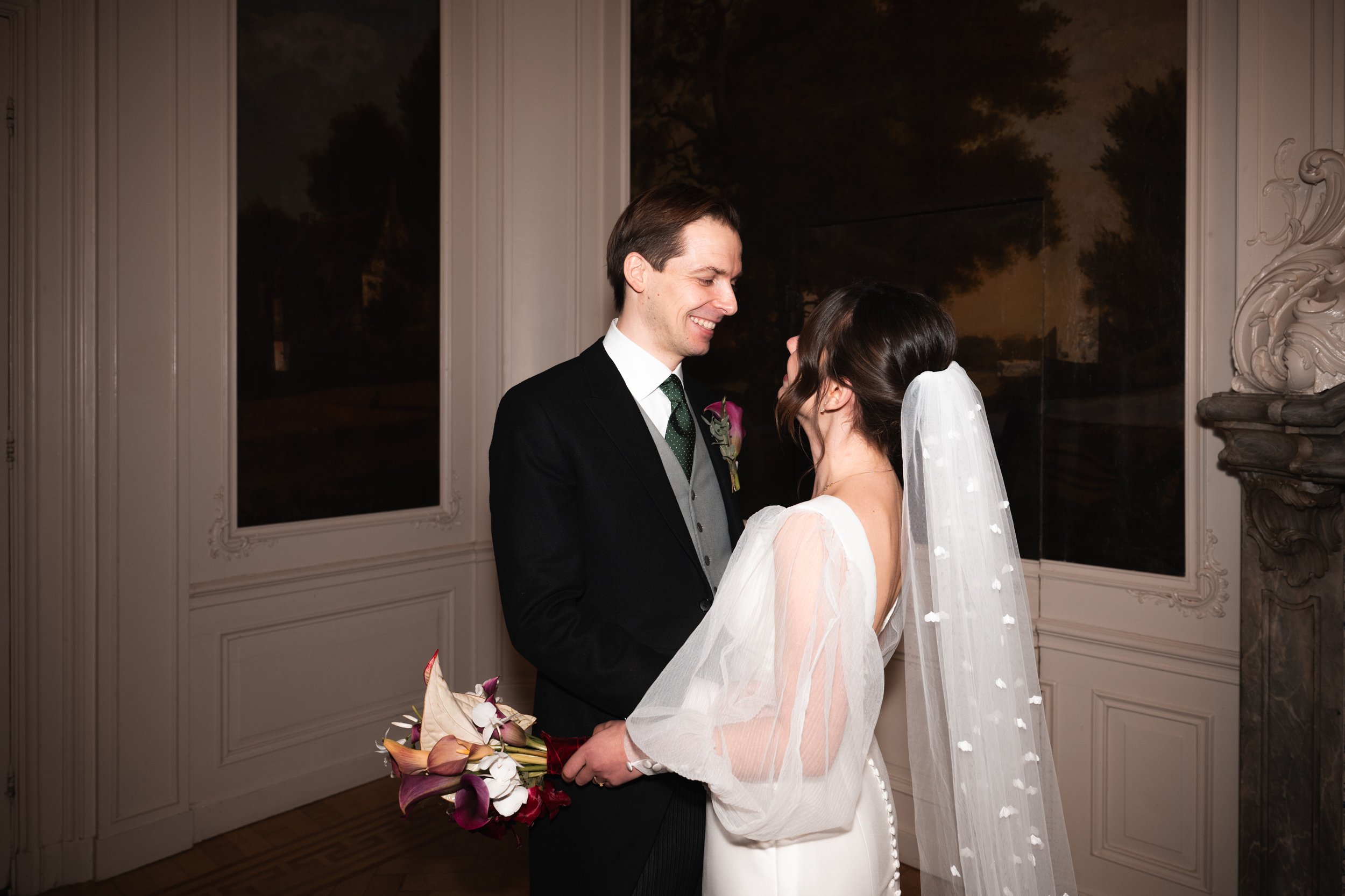 A bride and groom stand close together during their wedding ceremony indoors, smiling at each other. The groom is wearing a black suit with a white shirt and a green tie, holding a bouquet of flowers. The bride is wearing a white wedding dress and ve