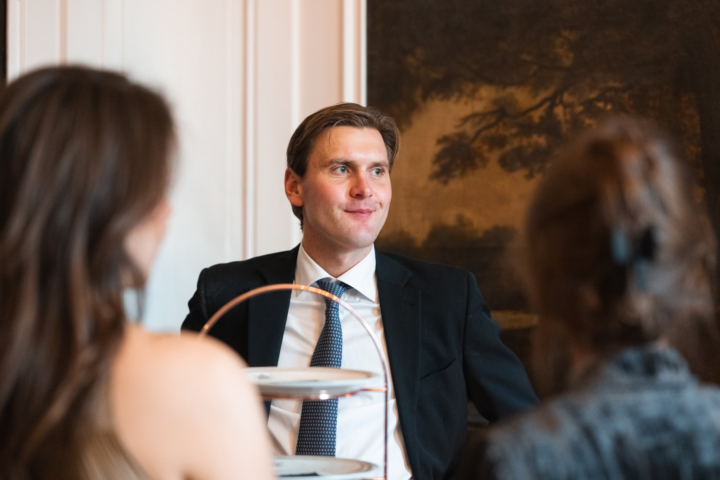 A man in a suit and tie sitting at a table with two women, in a room with wood paneling and a landscape painting in the background.
